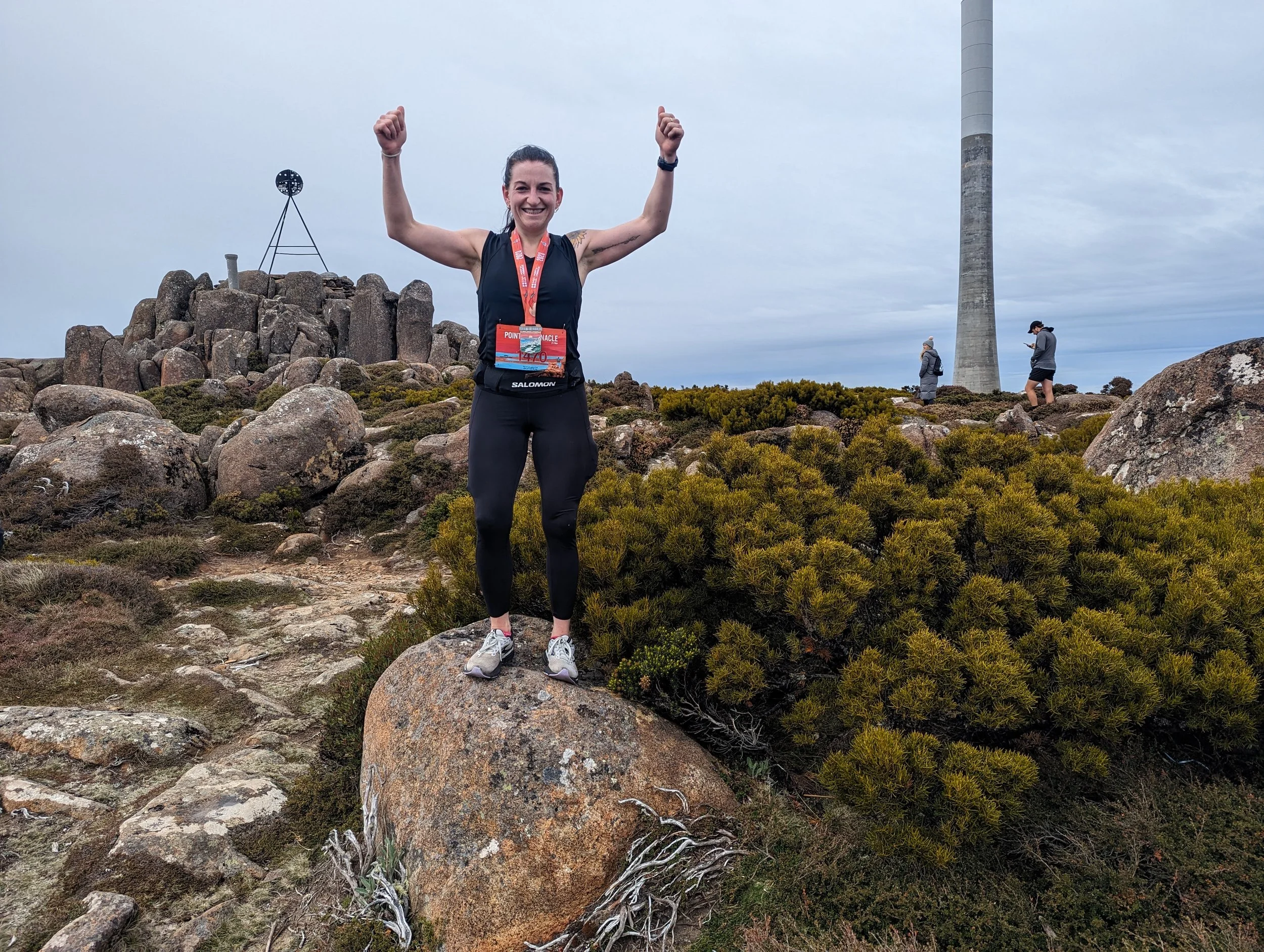 Smiling woman with a medal around her neck, celebrating at the top of a rocky hill with a tower and tall chimney in the background.
