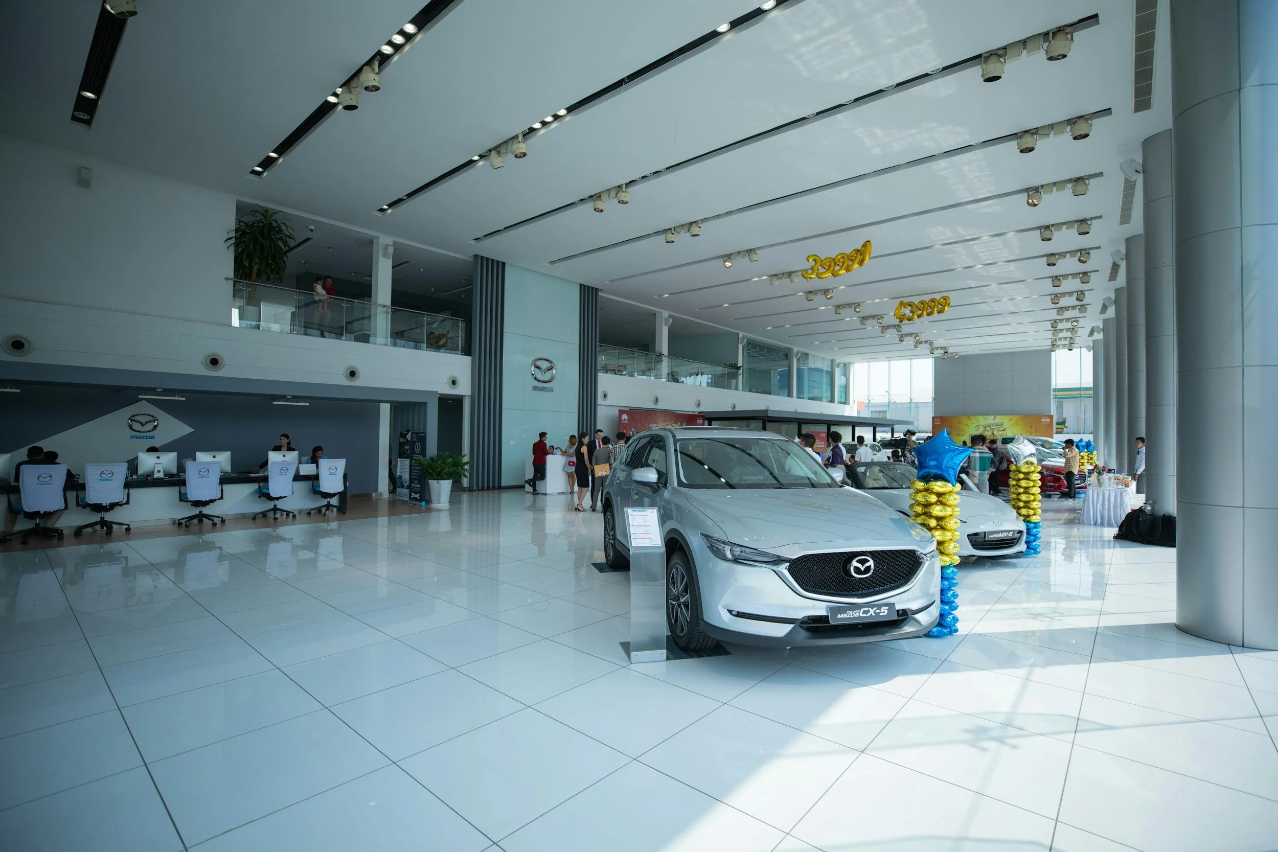 Inside a Mazda car dealership showroom with two Mazda vehicles on display, including a silver Mazda CX-5 in the foreground. There are balloons and decorations indicating a promotional event, and people are seen walking and sitting at a reception desk.