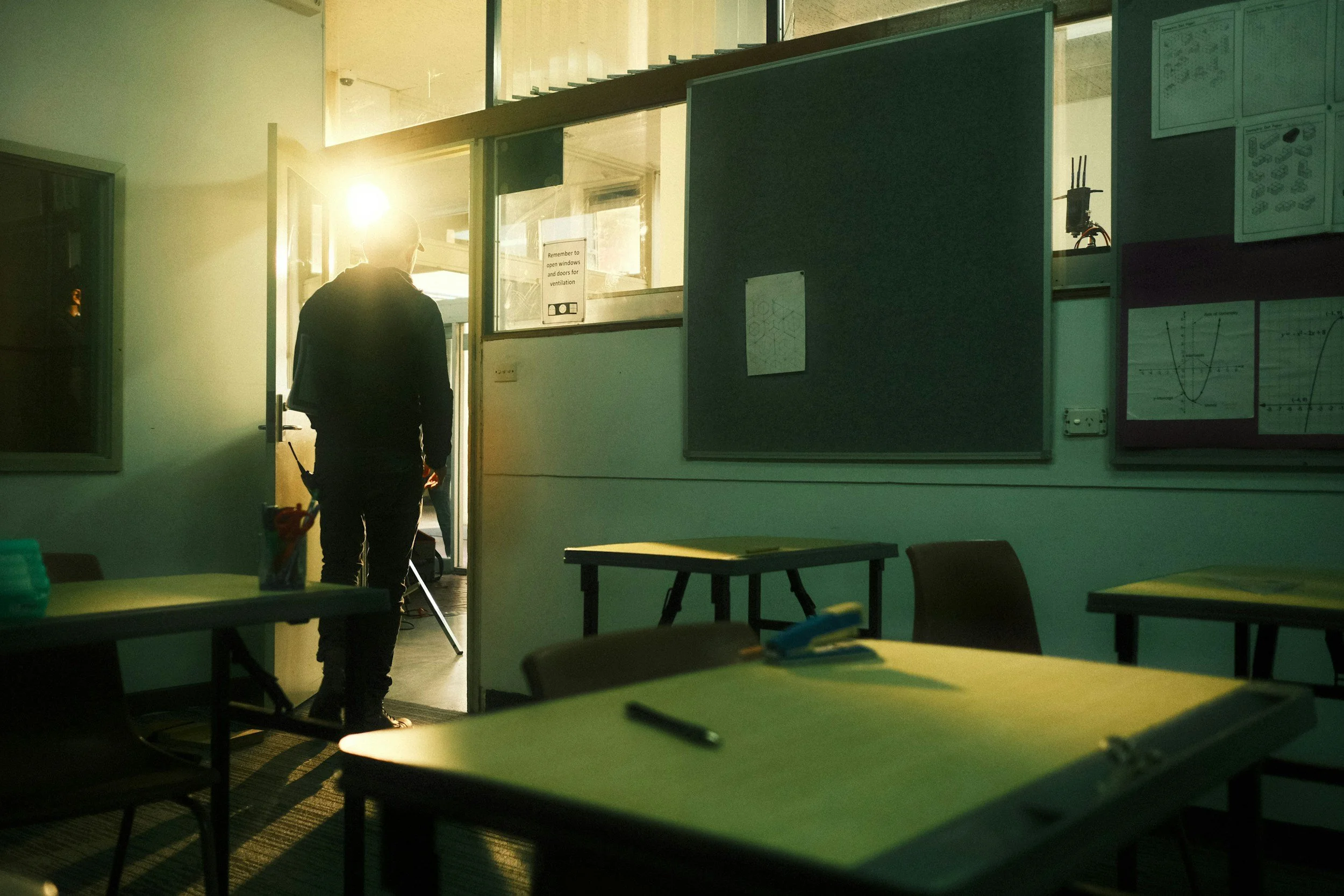 A person stands in a classroom or office space with sunlight streaming through the window, wearing glasses and dark clothing, with some desks and charts on the walls.