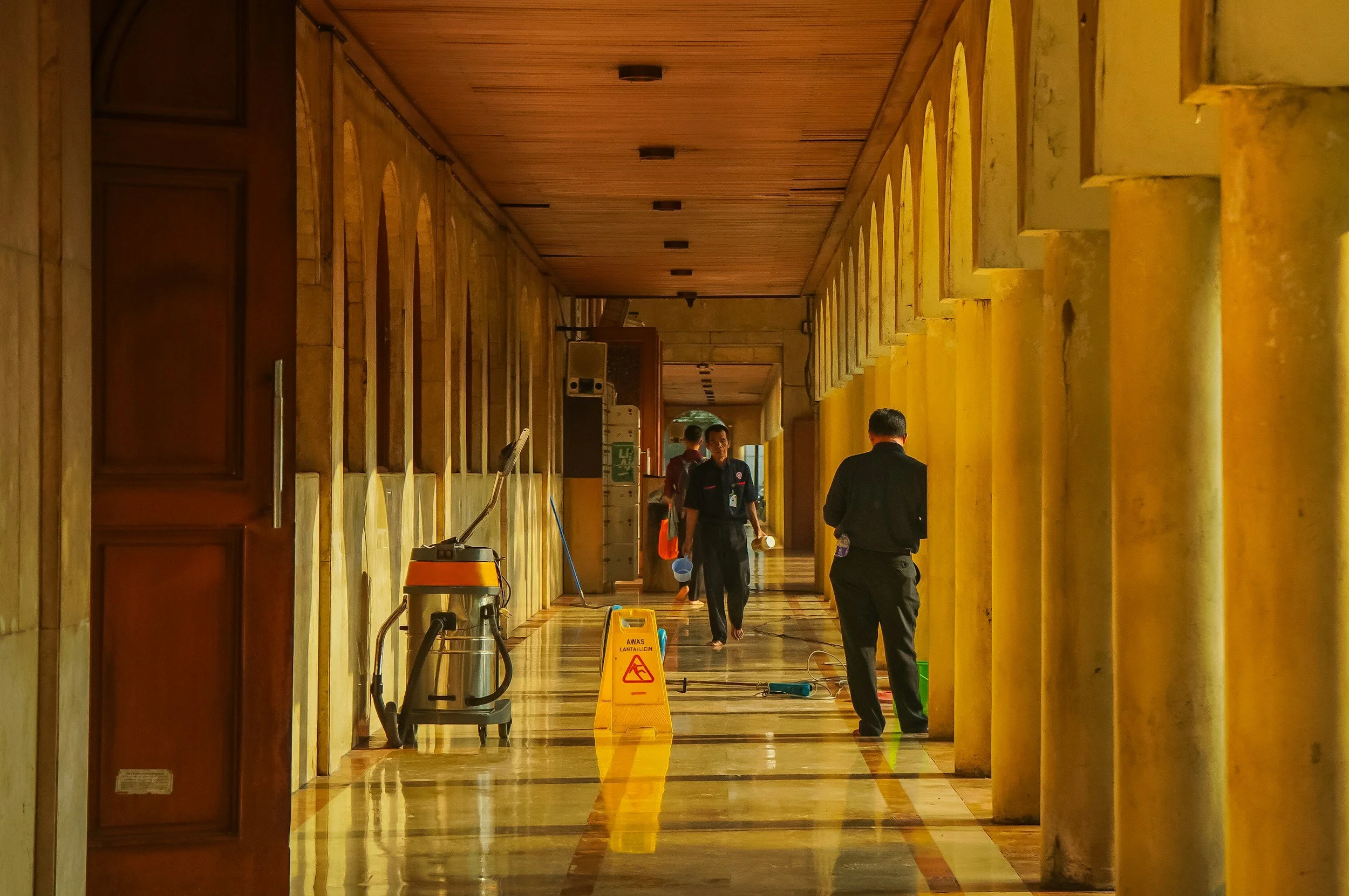 People walking through a brightly lit hallway under renovation or cleaning, with cleaning equipment and yellow caution signs visible.