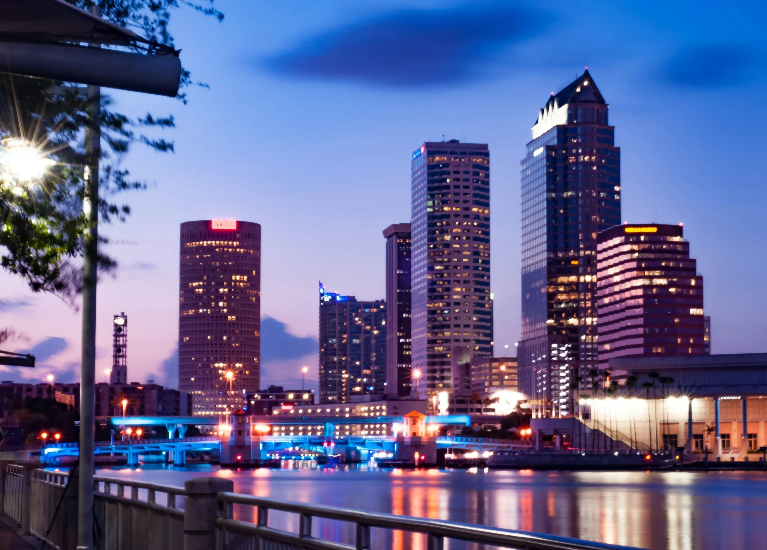 City skyline at dusk with tall modern buildings illuminated, a river with reflections, and a waterfront promenade with lights.