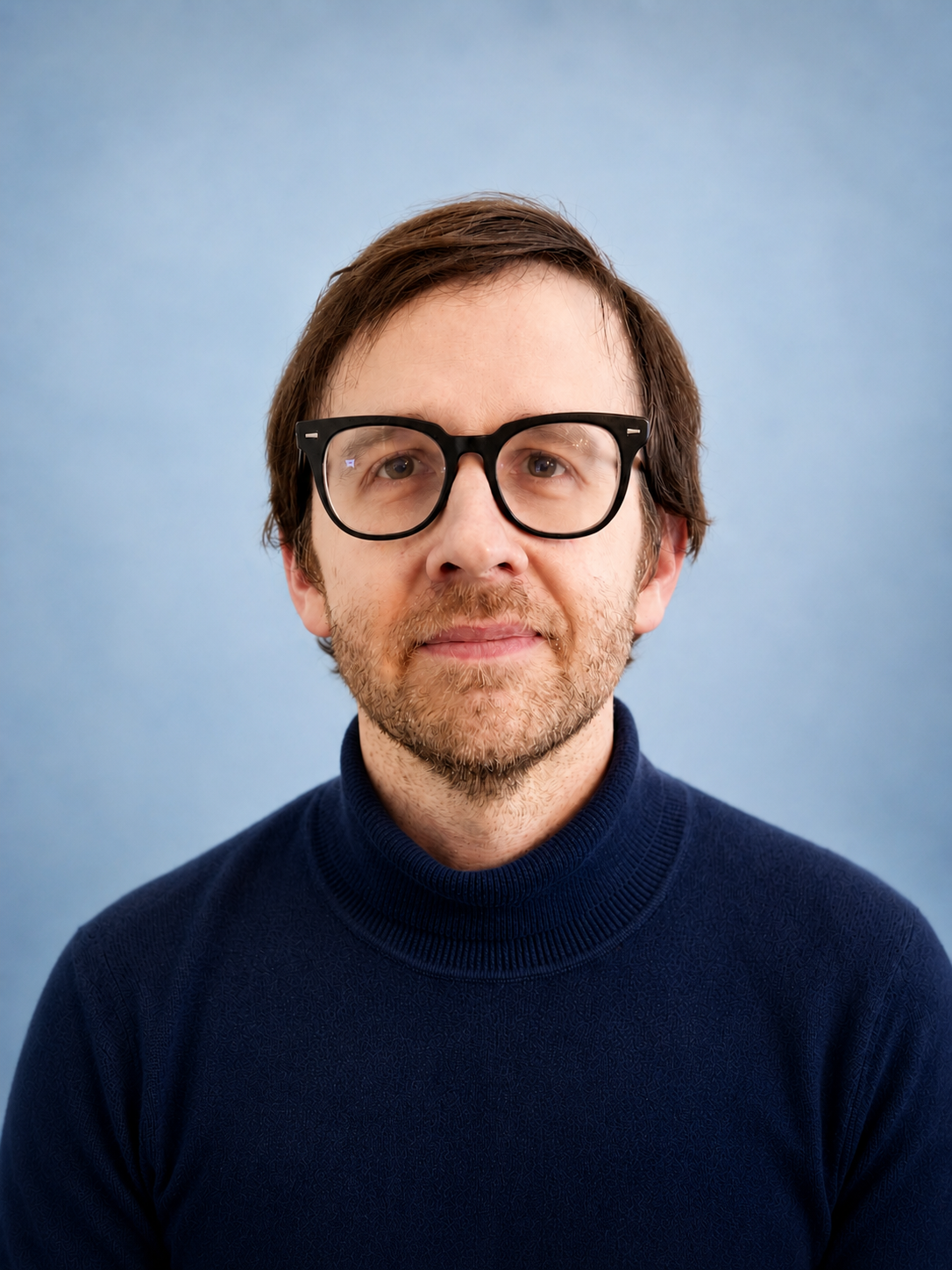 A man with brown hair, beard, and glasses, wearing a navy blue turtleneck sweater, against a light blue background.