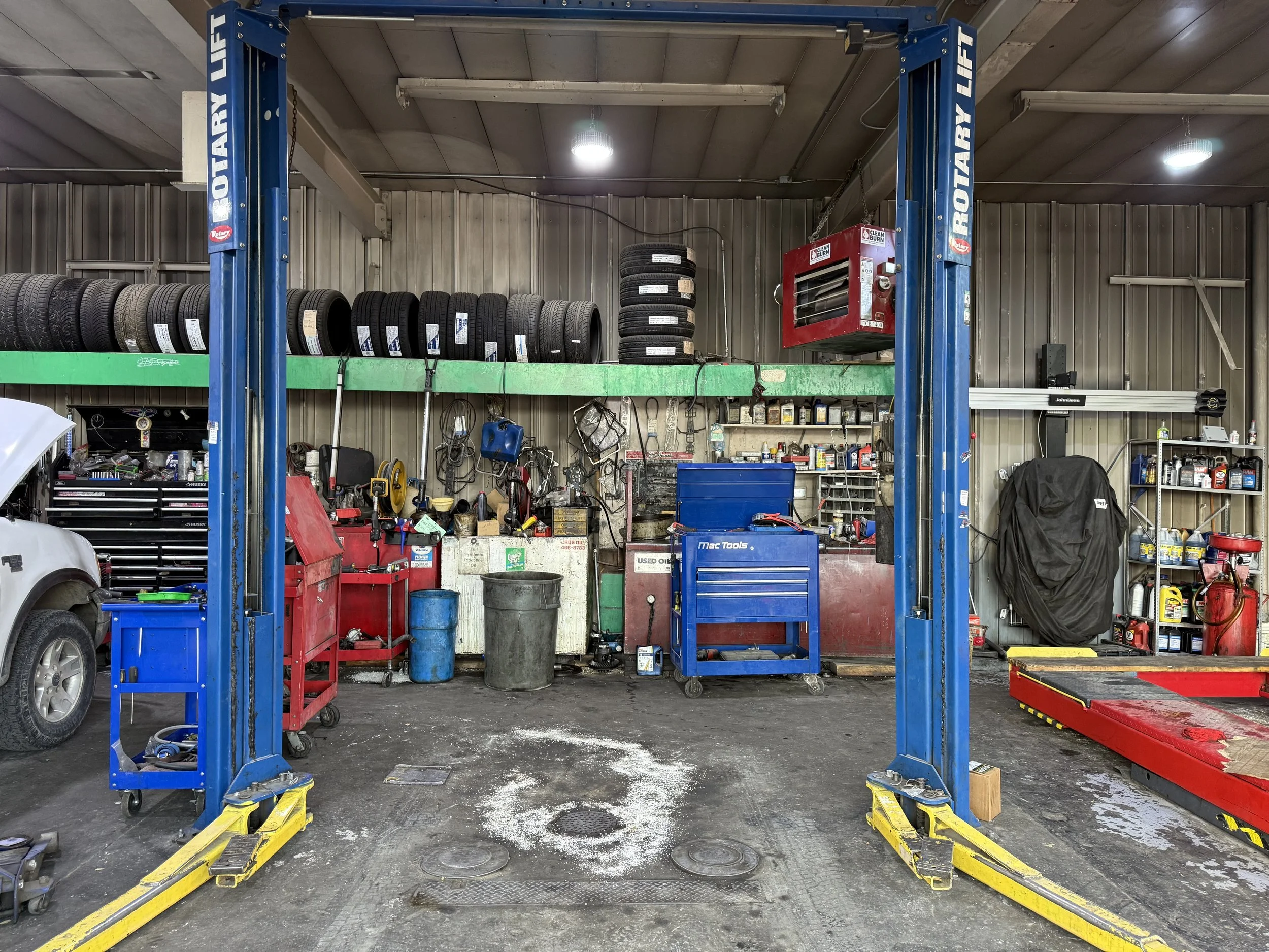 A garage workshop with a blue vehicle lift, tire stacks on a green shelf, cabinets, and various tools and equipment.