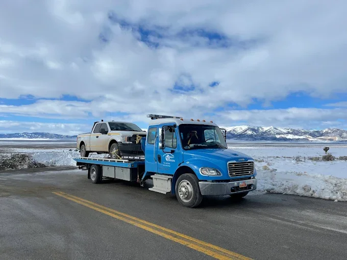 A blue tow truck driving on a snowy mountain road with a beige pickup truck on its flatbed.
