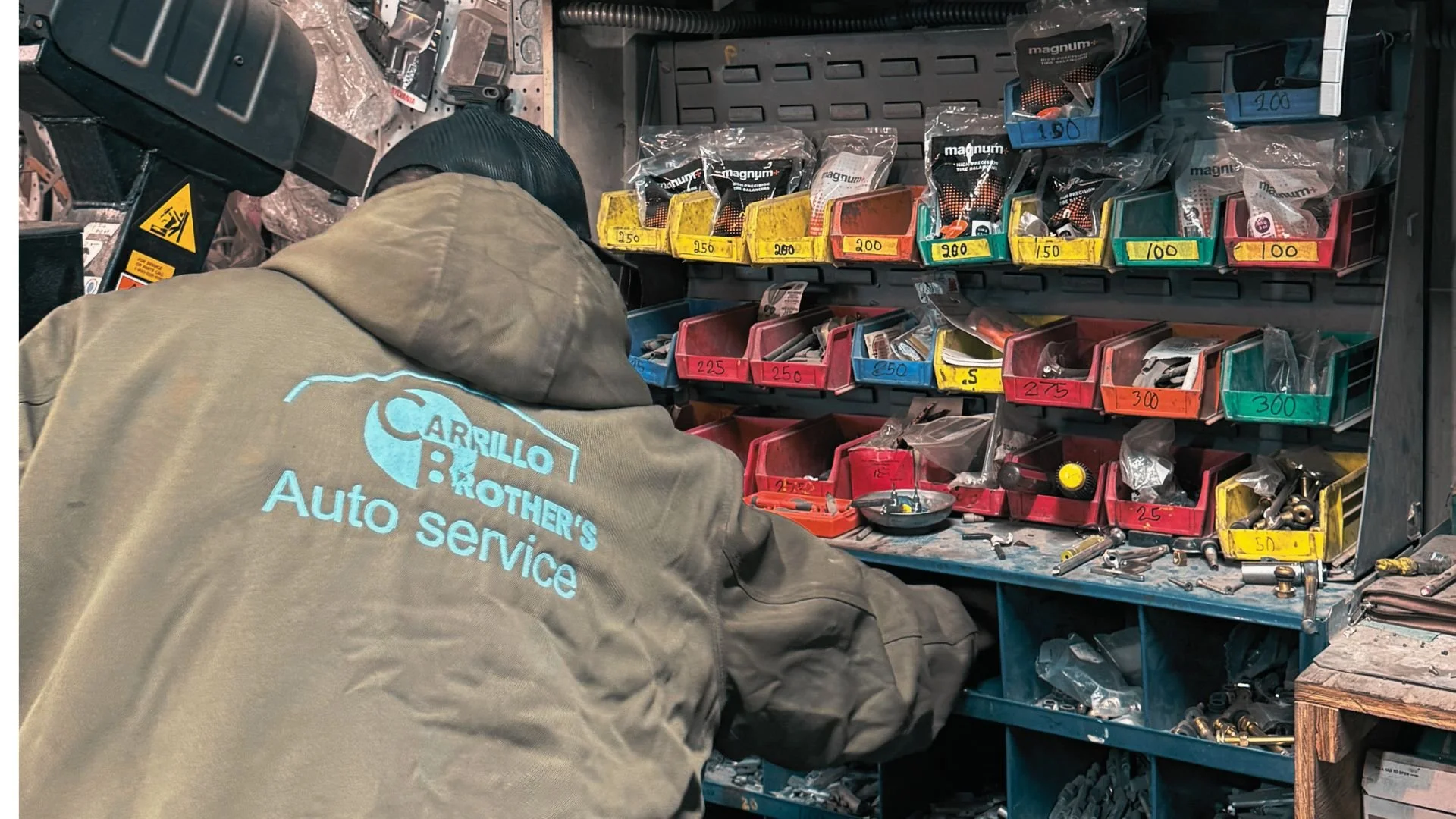 A worker at an auto service shop seen from behind, wearing a gray hoodie with the words 'Carrillo Brothers Auto Service' on the back. The person is sorting through various small automotive parts and tools organized in compartments on a workbench, which also has loose parts and containers.