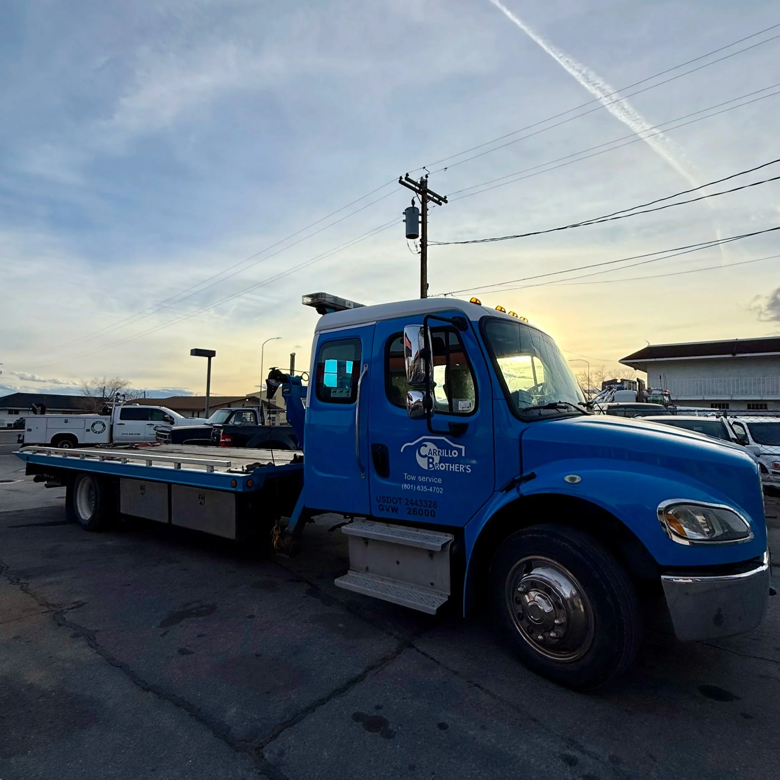 A blue tow truck labeled Carrillo Brothers Tow Service is parked in a lot with other vehicles. The sky shows wispy clouds and contrails from airplanes at sunset.