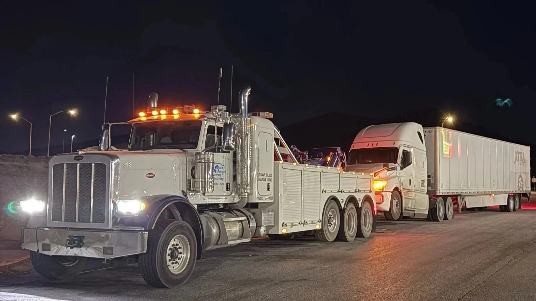 A large white semi-truck with multiple axles and yellow lights on the cab, hauling a long white trailer, parked at night with streetlights nearby.