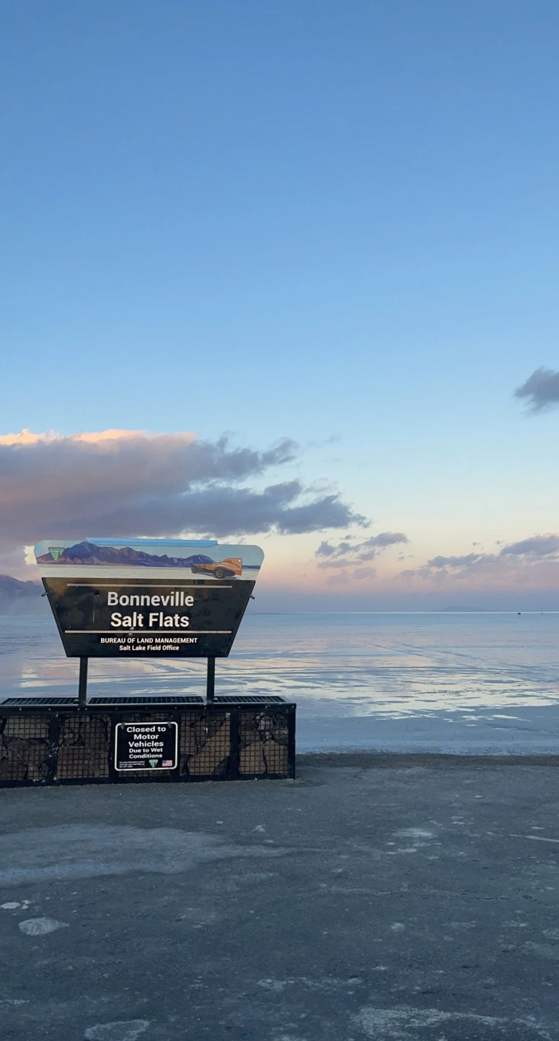 A sign for Bonneville Salt Flats in Utah, with a background of mountains and a body of water, under a sky with clouds and a setting or rising sun. There is a sign below that says the area is closed to motor vehicles due to wet conditions.