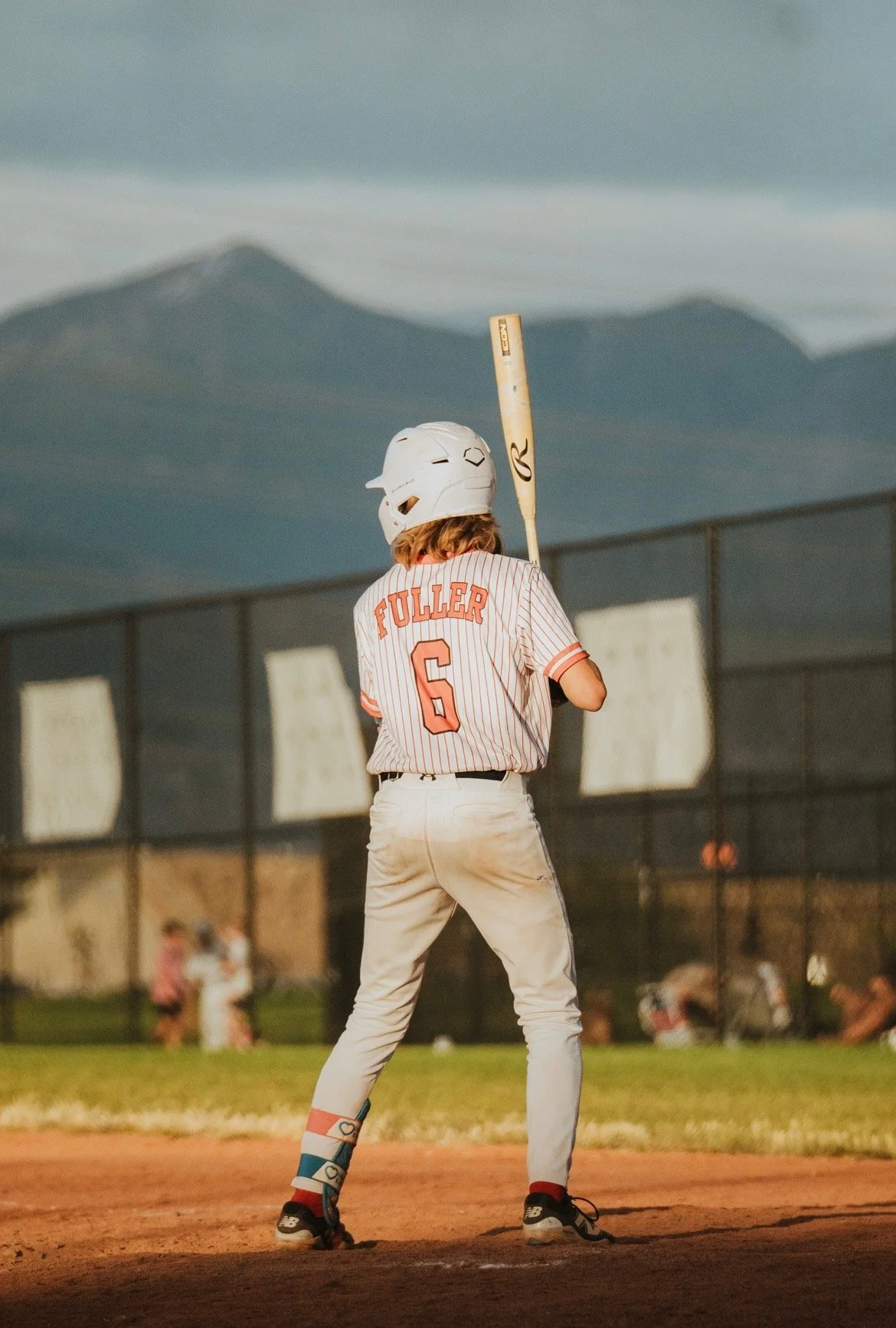 A young baseball player wearing a striped jersey with 'Fuller' and the number 6 on the back, standing on a baseball field about to bat, with mountains and a chain-link fence in the background during sunset.