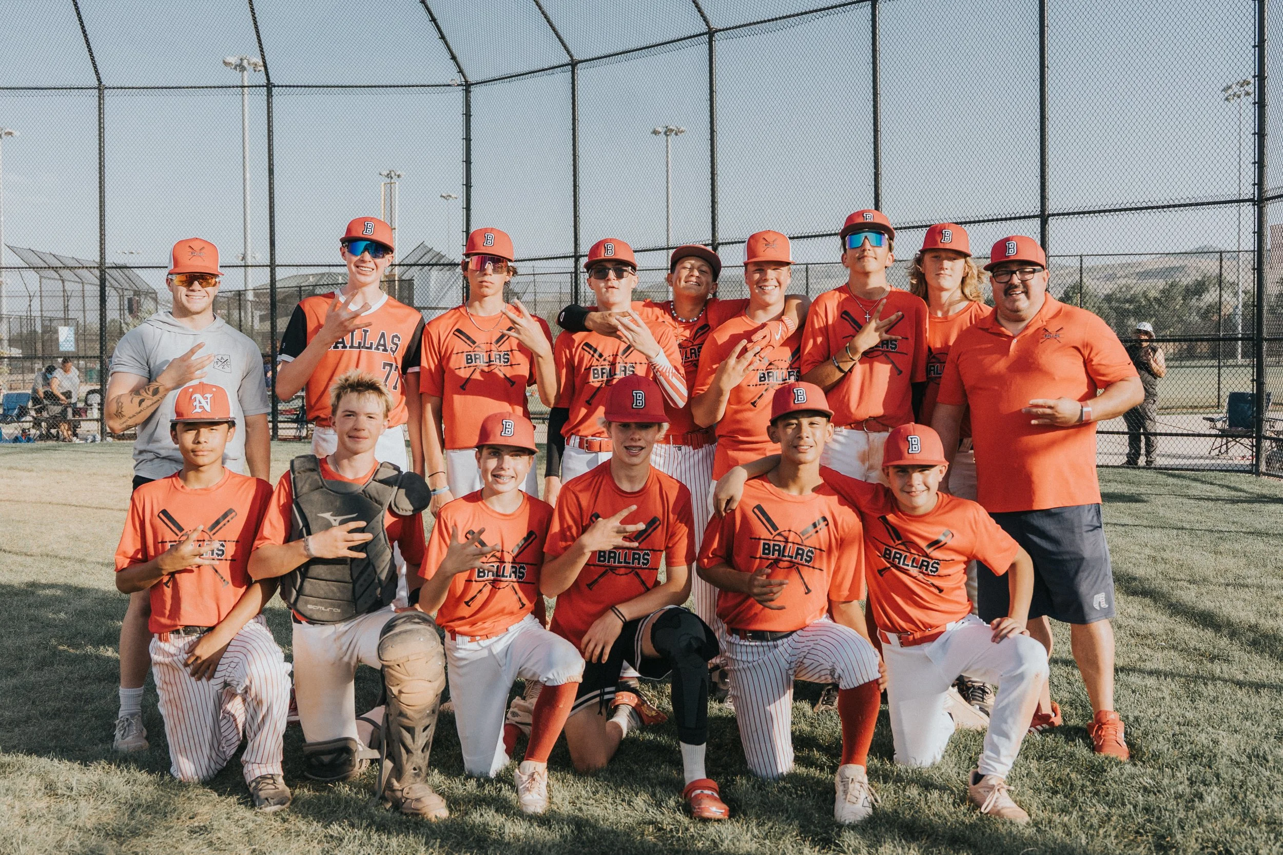 A youth baseball team with coaches and players posing on a baseball field, dressed in orange jerseys and caps, with a chain-link fence in the background.