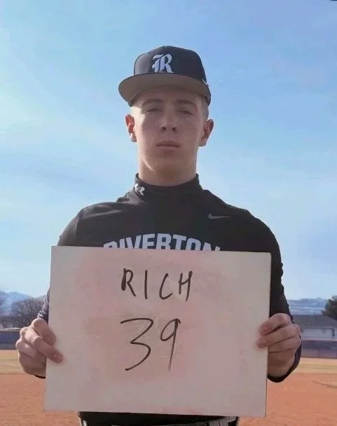 A young man wearing a black cap with a white 'R' and a black jacket holding a sign that reads 'RICH 39' outdoors on a sunny day.