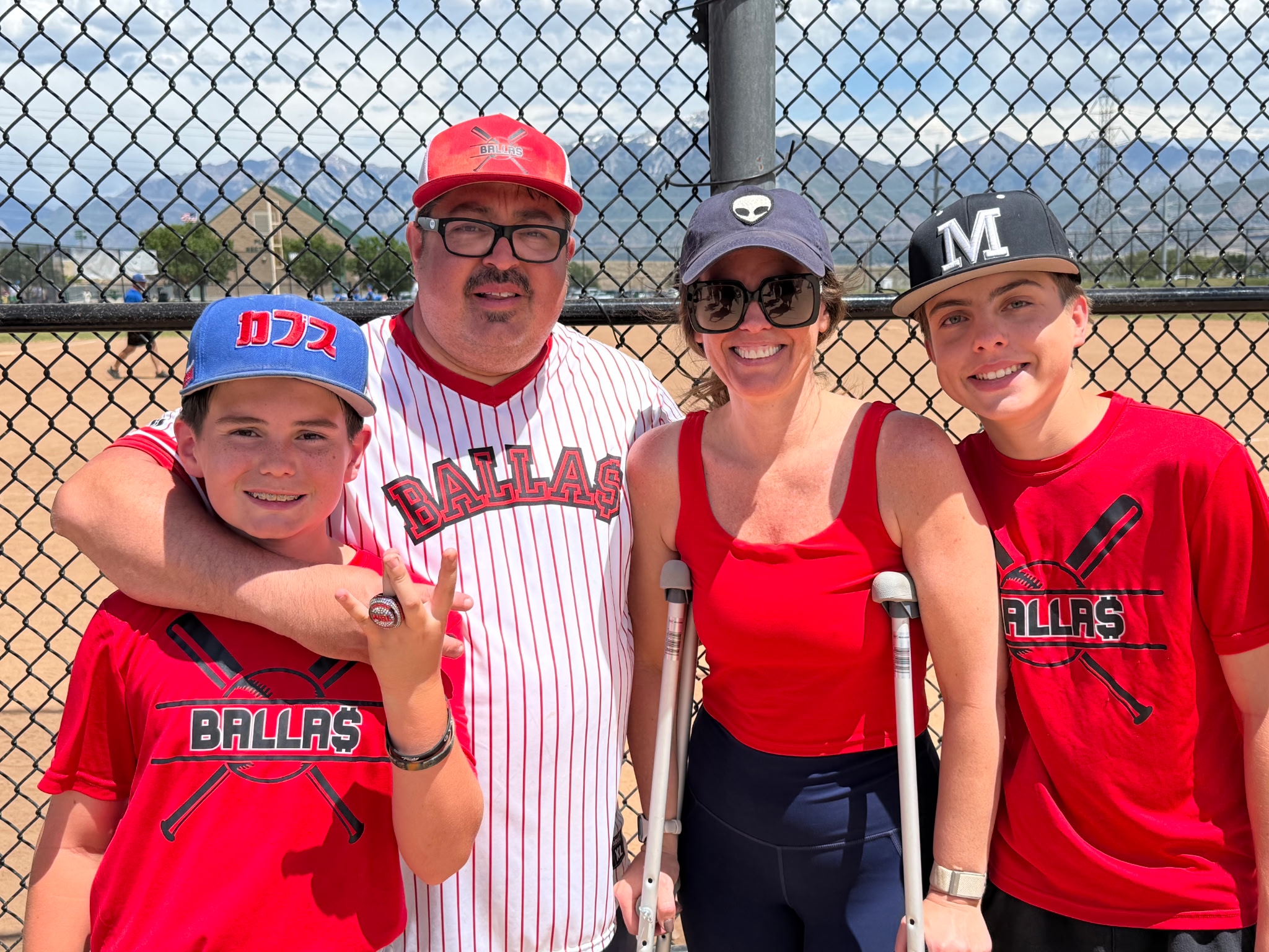 A group of four people, a man, woman, and two boys, standing in front of a chain-link fence at a baseball field. They are all smiling, dressed in red sportswear, with two of the boys wearing baseball caps. The background shows a park, some buildings, snow-capped mountains, and power lines.