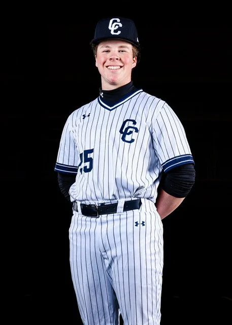 Young male baseball player in a white pinstripe uniform with navy accents, wearing a black cap with a white logo, standing against a black background, smiling with hands behind his back.