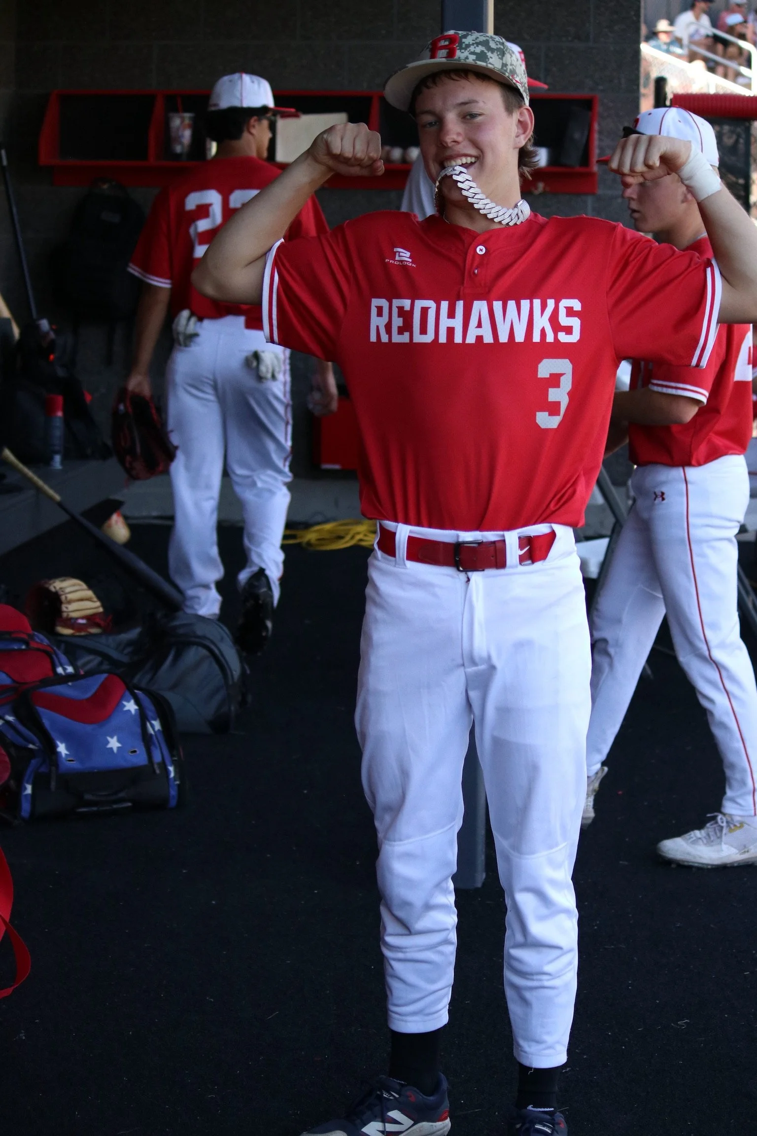A teenage baseball player wearing a red jersey with 'REDHAWKS' and the number 3, flexing his arms and smiling, inside a dugout with other players and baseball equipment.