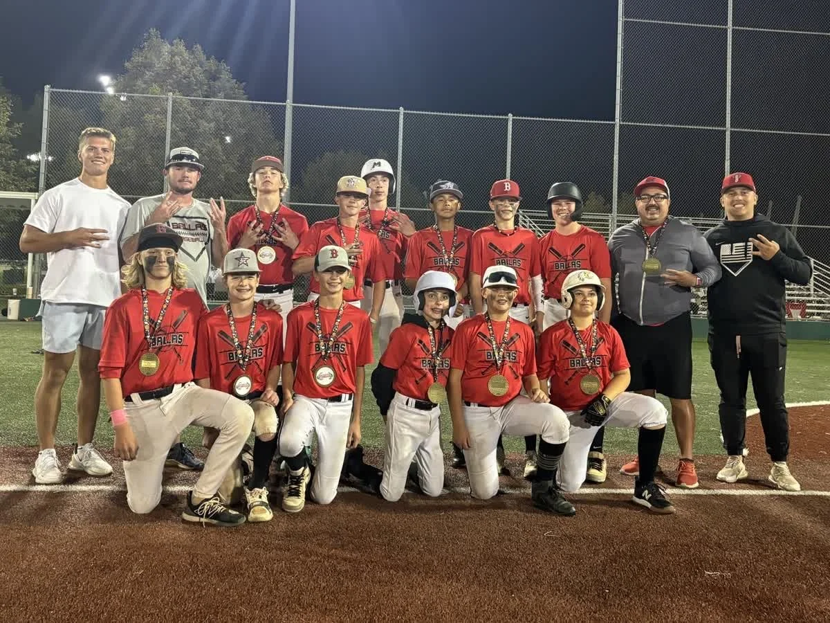 Youth baseball team and coaches posing for a photo on a baseball field at night, with team members wearing red jerseys and medals around their necks.
