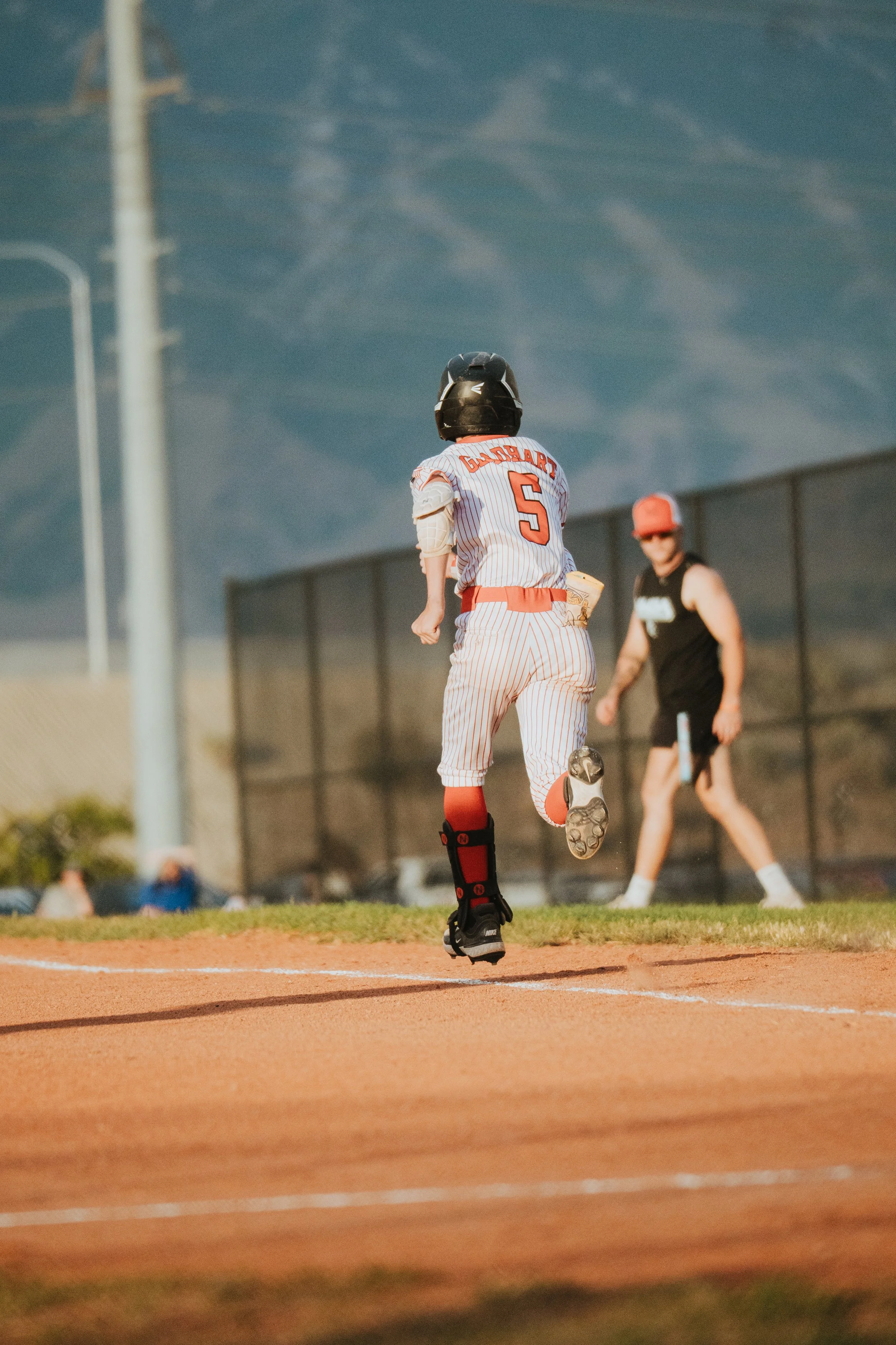 A young baseball player wearing a helmet and jersey with the number 5 on it runs on a baseball field, with a person wearing a tank top and cap in the background and a dark cloudy sky overhead.