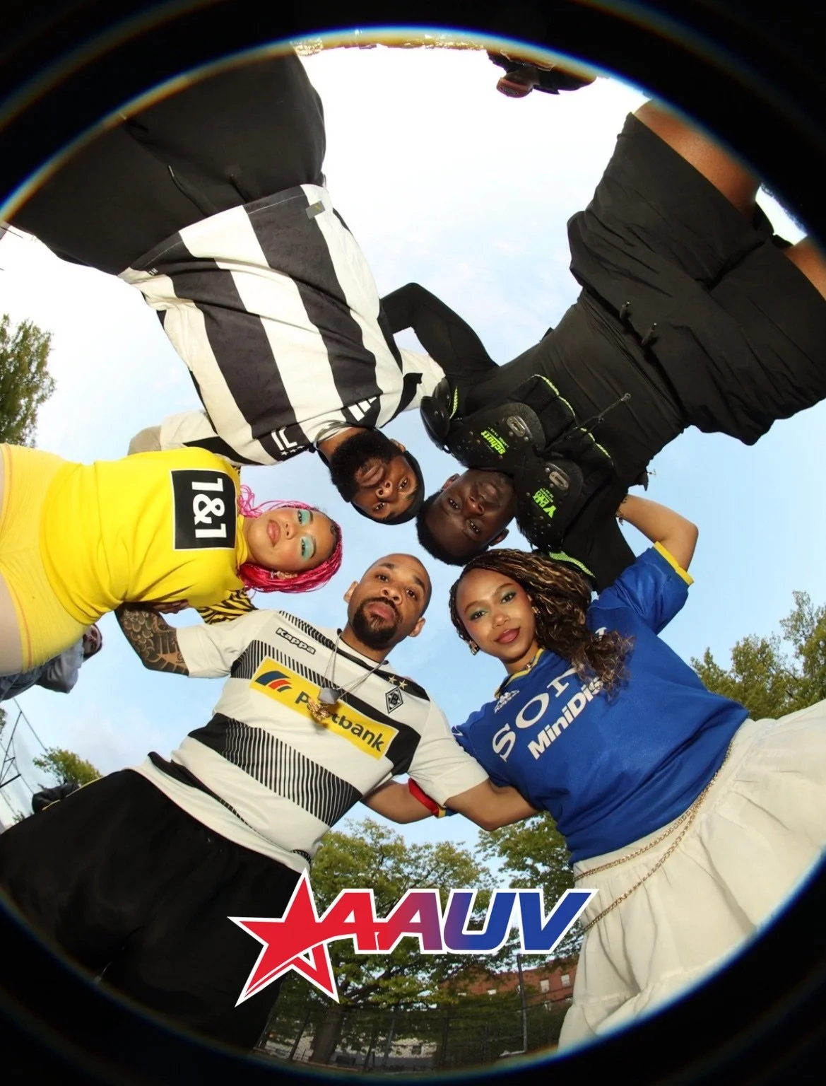 A group of six diverse people standing in a circle outdoors, looking down at the camera which is positioned on the ground. The sky and trees are visible in the background.