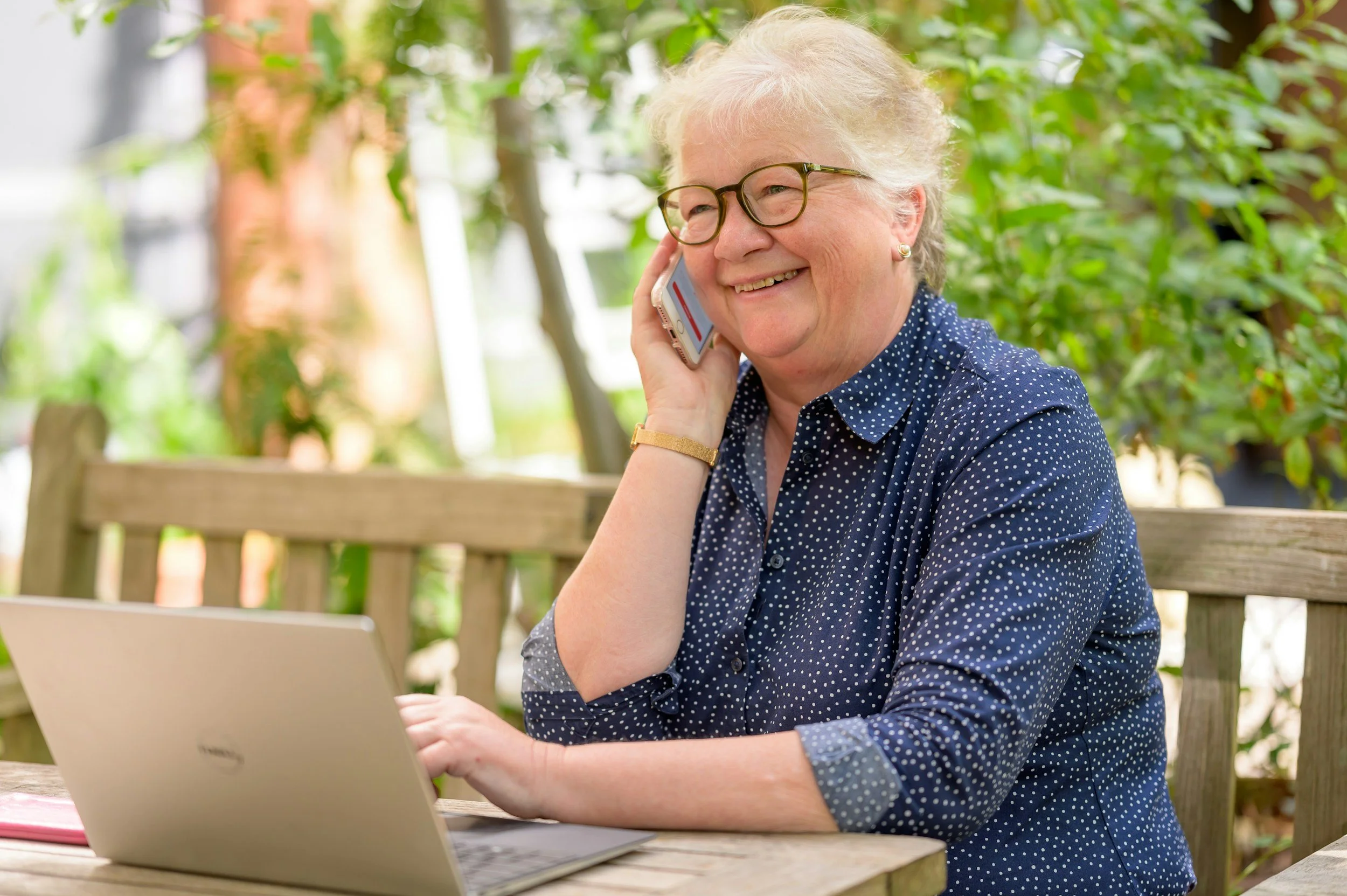 Smiling elderly woman with gray hair and glasses talking on her cell phone while sitting at an outdoor wooden table with a laptop. Patient Care Advocacy. Florida. Healthcare Navigation Services. Insurance Navigation Services.