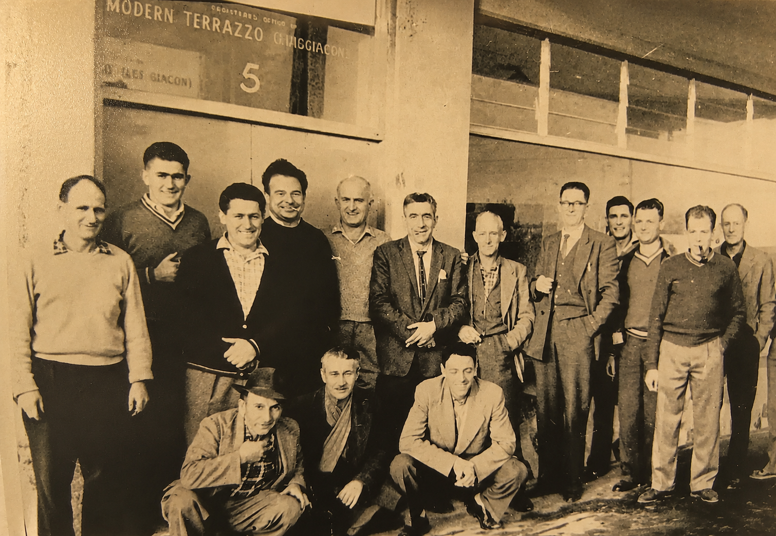 Black and white historical photo of fifteen men posing together outside a building with sign reading "Modern Terrazzo G. Migliaccio." The men are dressed in suits, sweaters, and casual jackets, some wearing ties, in a vintage mid-20th-century style.