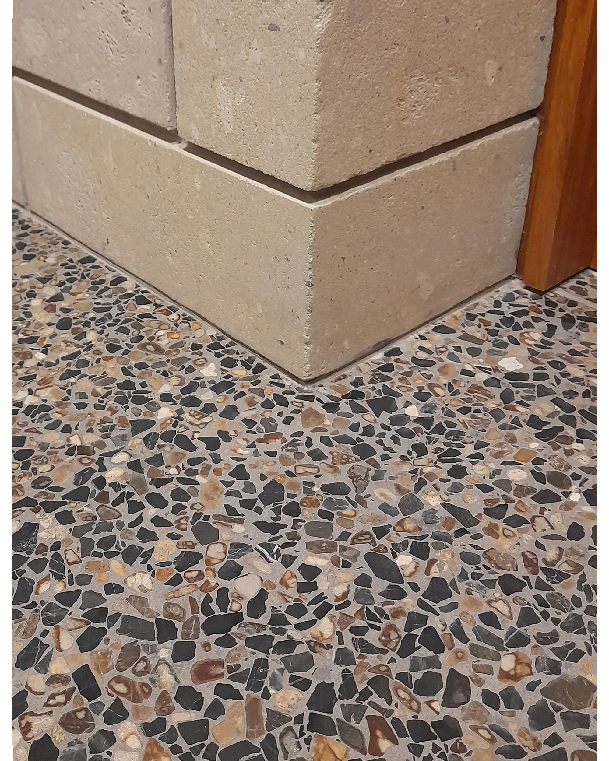 Corner of a tiled wall above a terrazzo floor, with a wooden trim on the right.