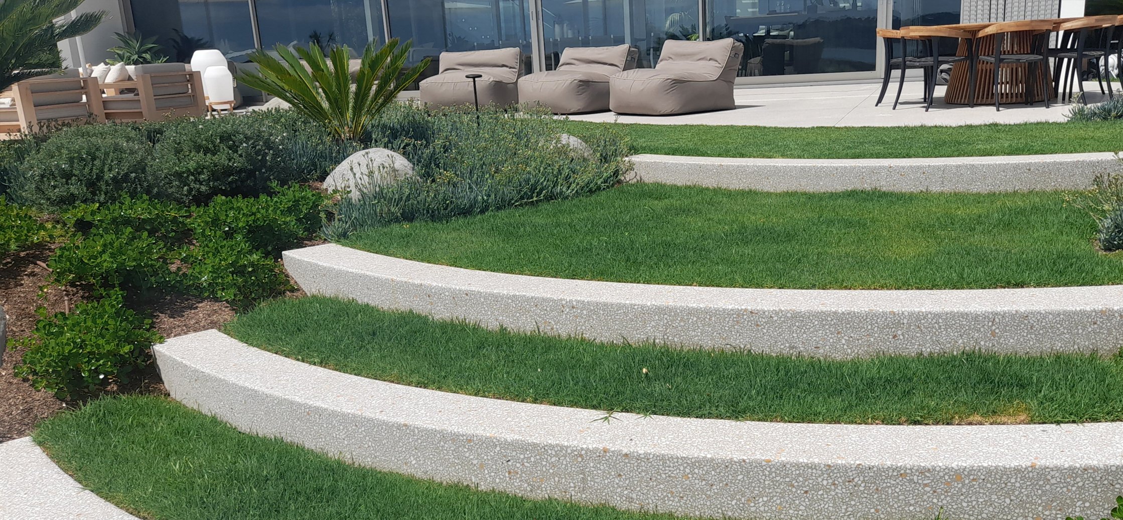 A modern rooftop terrace with planted greenery, curved white concrete steps, and outdoor seating including beige cushioned chairs and a wooden dining table with black chairs.
