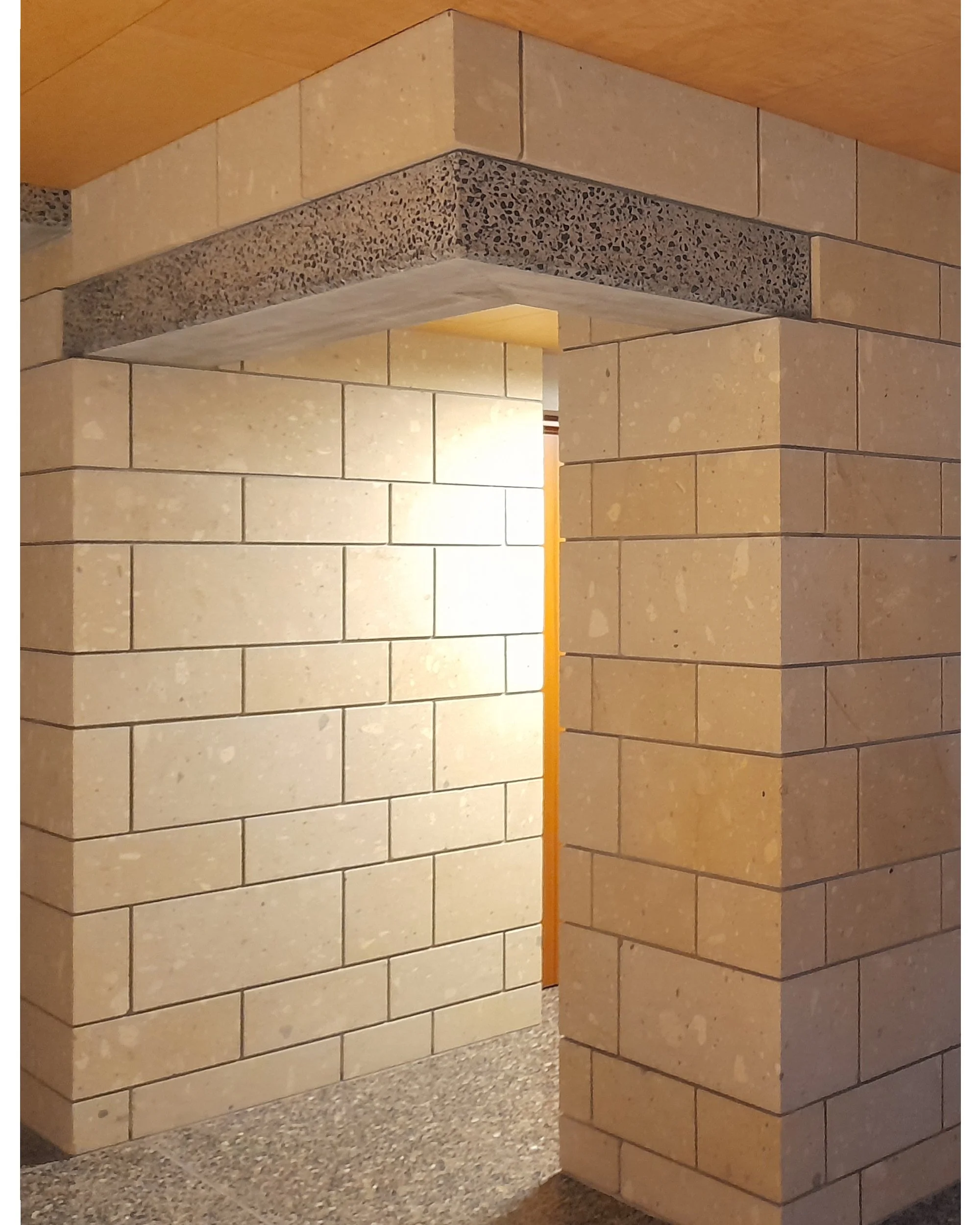 A corner of a room showing beige stone brick walls and a wooden ceiling with an open archway leading to another room.
