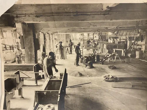 Black and white photo of factory workers assembling furniture on a factory floor with various tools and machinery.