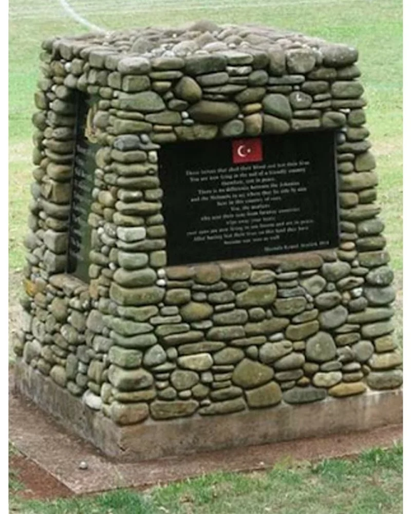 A stone monument with a black plaque, featuring a Turkish flag symbol, located outdoors on grass.