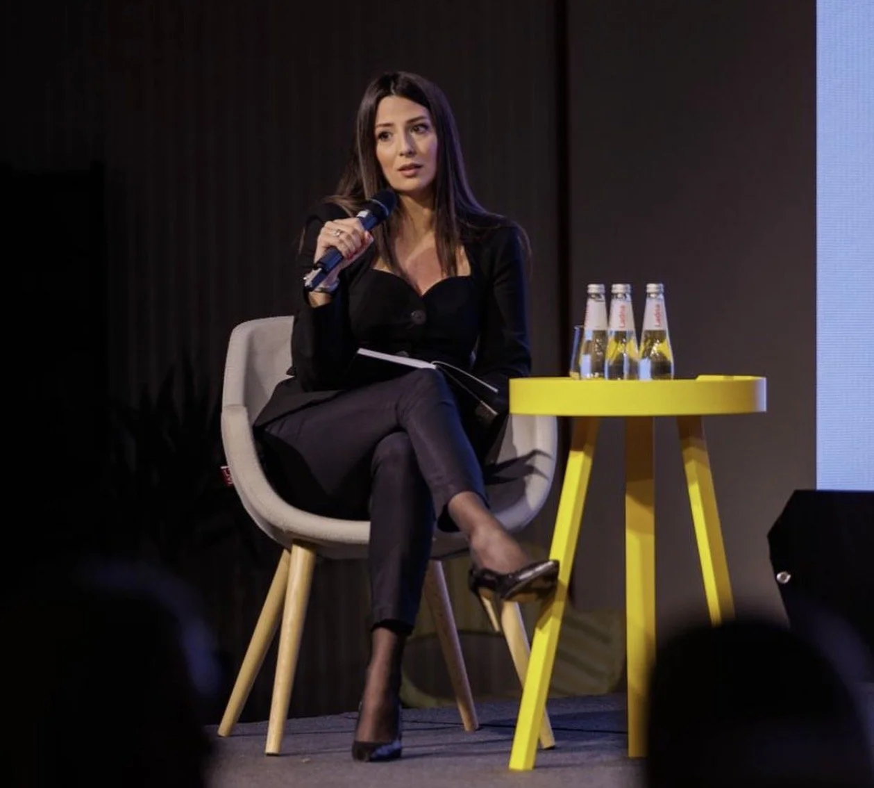 A woman with long dark hair in a black blazer and pants sitting in a cream chair, speaking into a microphone on stage. There is a yellow table with four bottled drinks on it beside her.