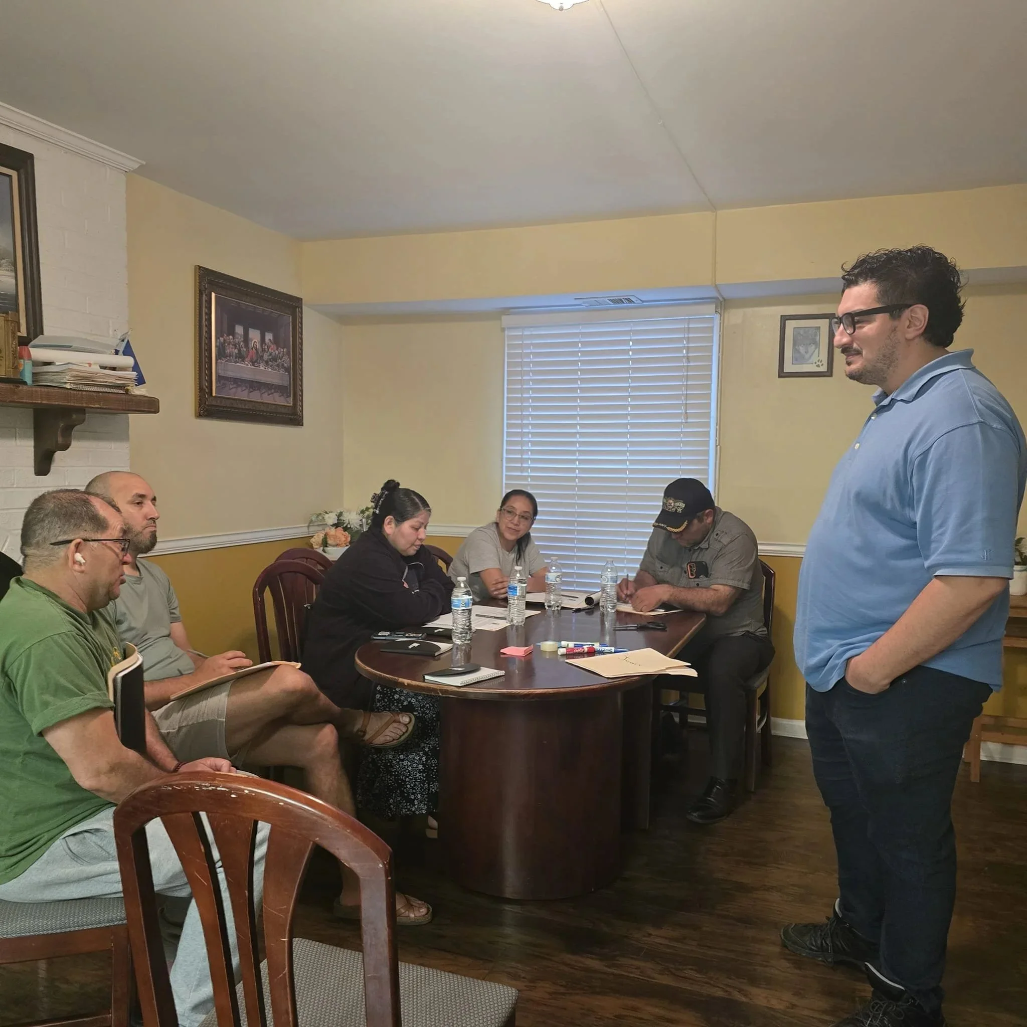 People sitting around a wooden table in a room with yellow walls, framed pictures, and a window with blinds. They are engaged in conversation, with water bottles and notebooks on the table. A man in a blue shirt is standing.