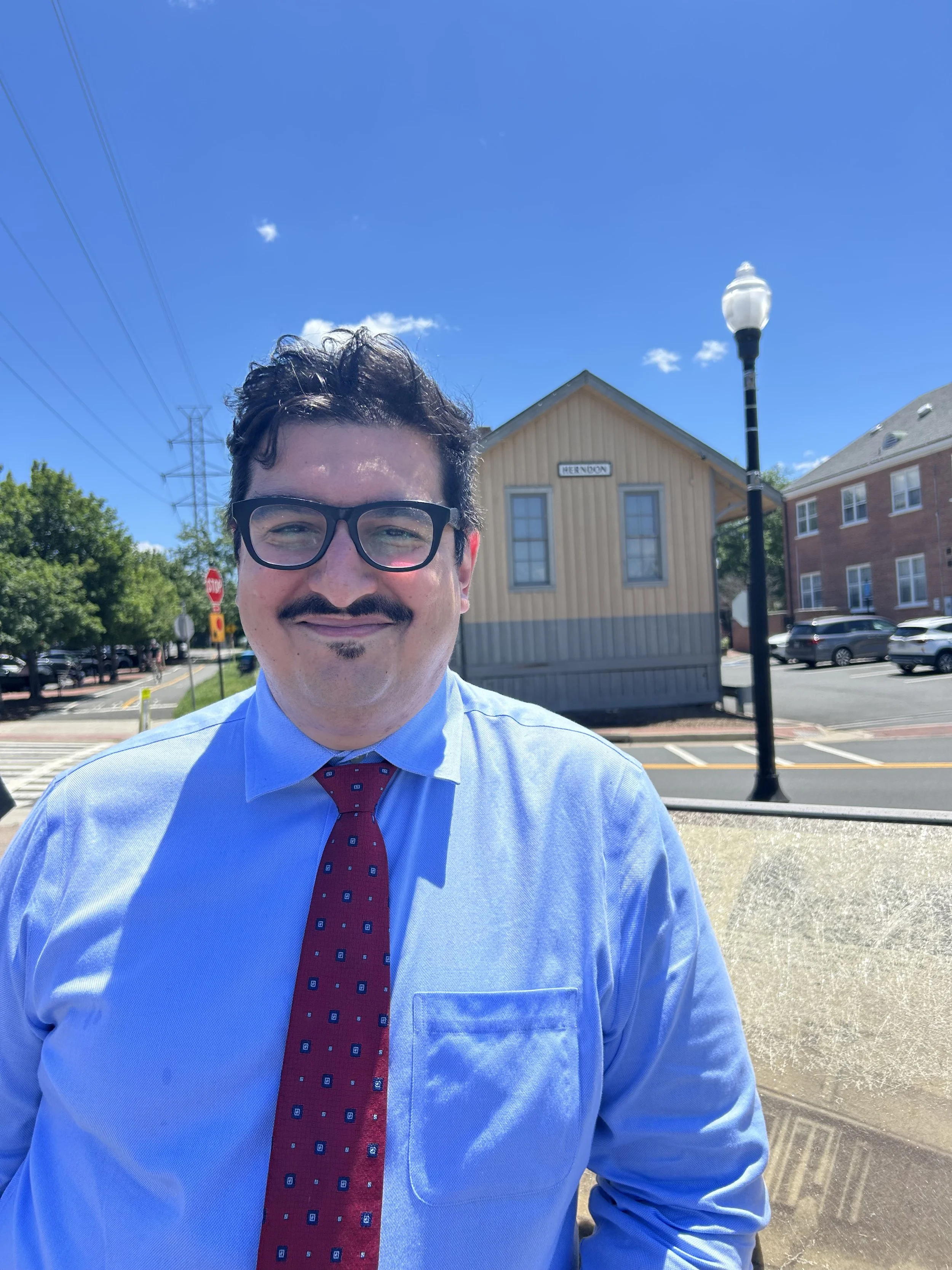 A man wearing glasses, a blue dress shirt, and a red tie takes a selfie outdoors on a sunny day. There are trees, a streetlamp, parked cars, and buildings in the background, including a small building with a sign that reads 'HERNDON'.
