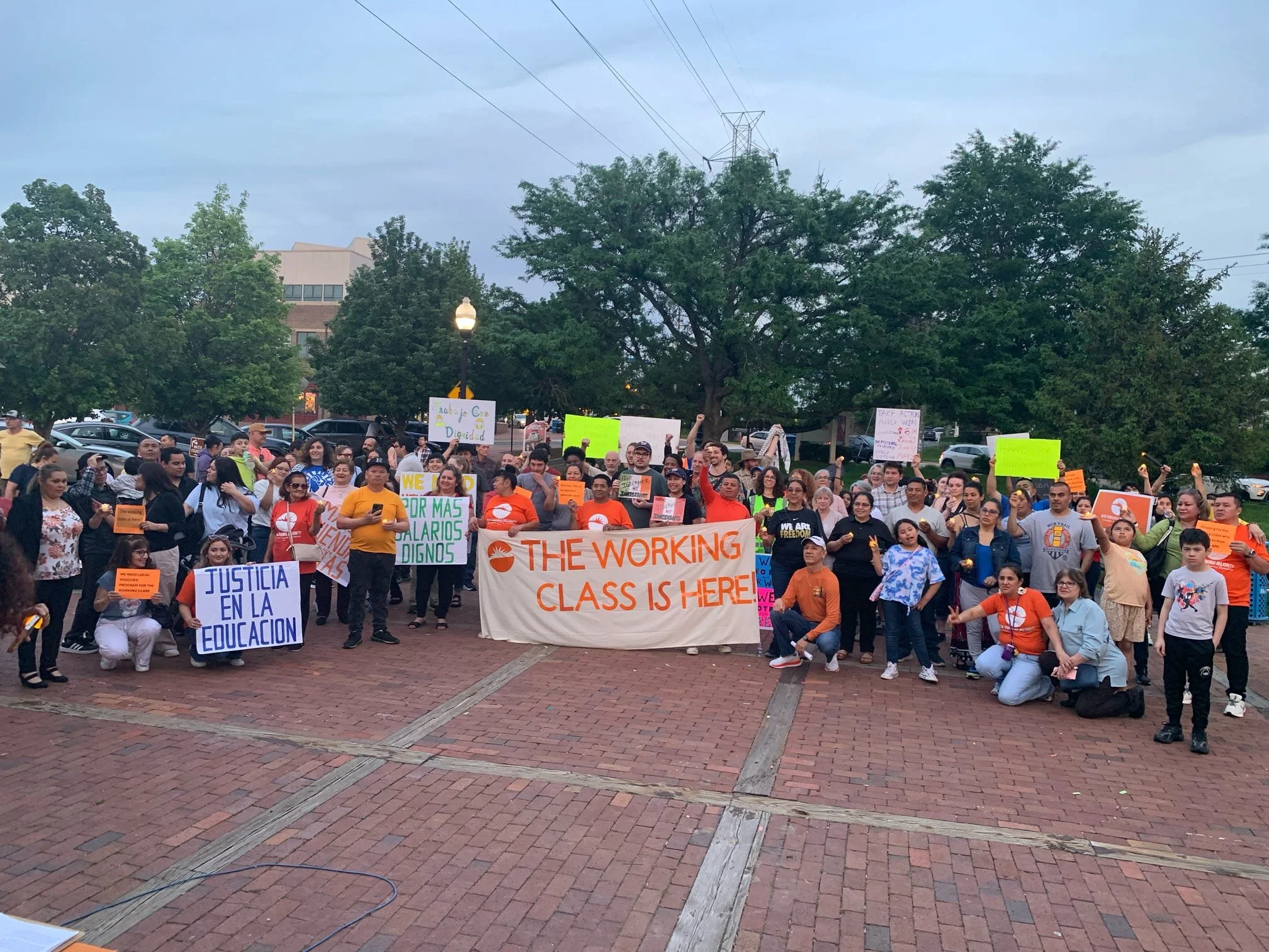 A large crowd of people protesting outdoors, holding colorful signs with messages advocating for education justice, fair wages, and workers' rights. The protest is set in a park or plaza area with trees, a lamppost, and a building in the background.