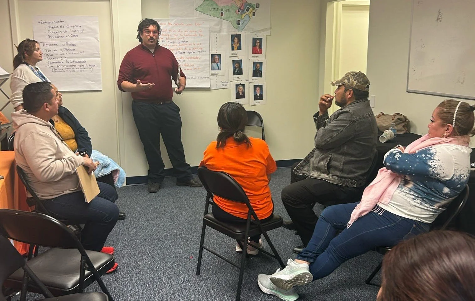 A man with glasses and a red shirt is standing and speaking to a small group seated in a room with charts and photographs on the walls.