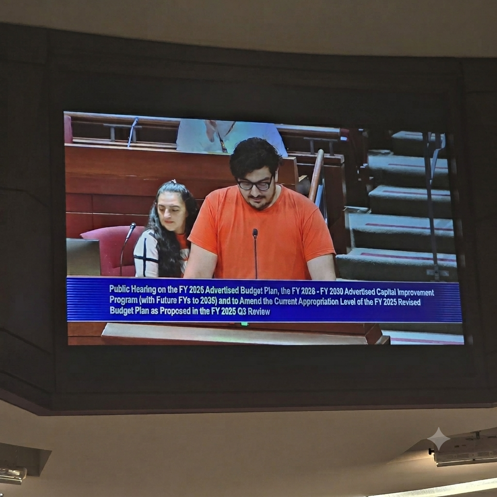 A man in an orange shirt speaking at a public hearing, with a woman sitting behind him, in a formal setting with multiple levels of seating.