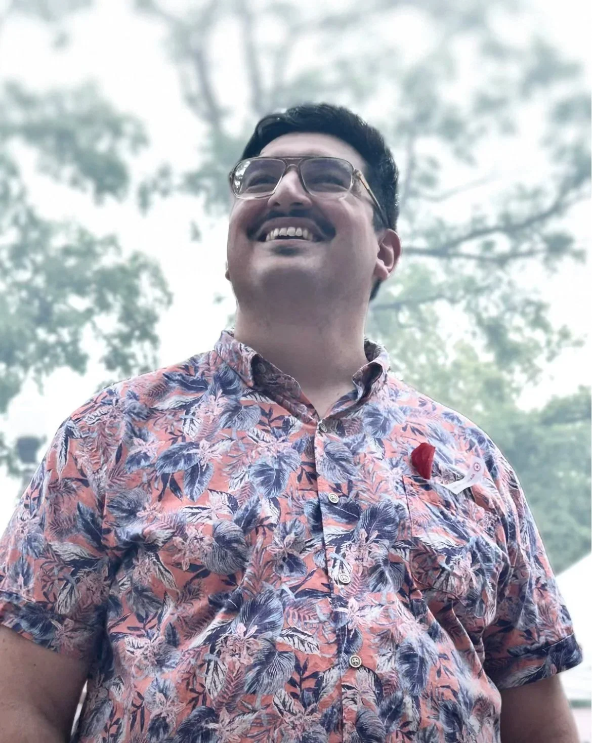 A man wearing glasses and a floral button-up shirt, smiling outdoors with trees and cloudy sky in the background.