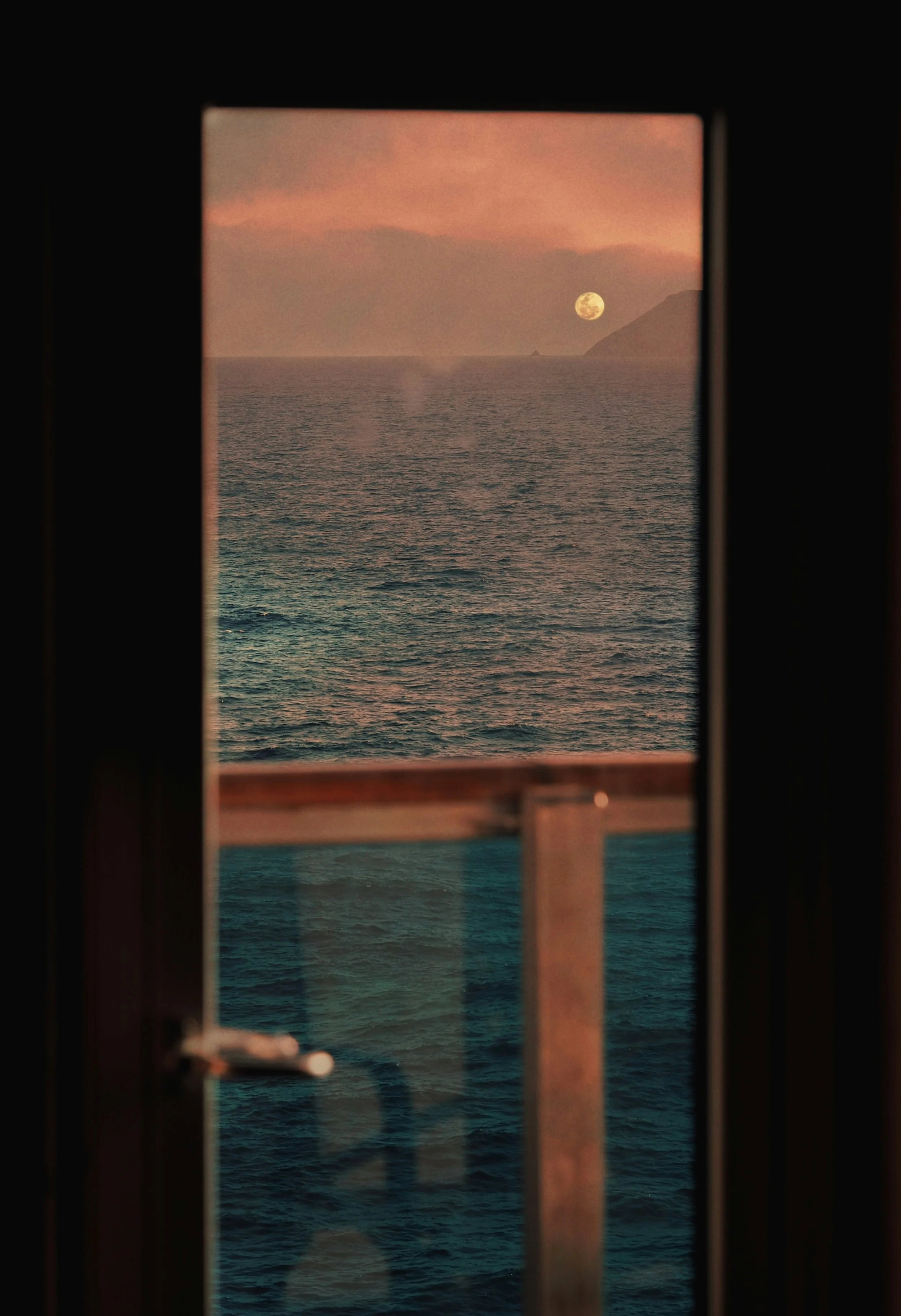 Sunset over the ocean with the moon visible, seen through a window with a dark frame.