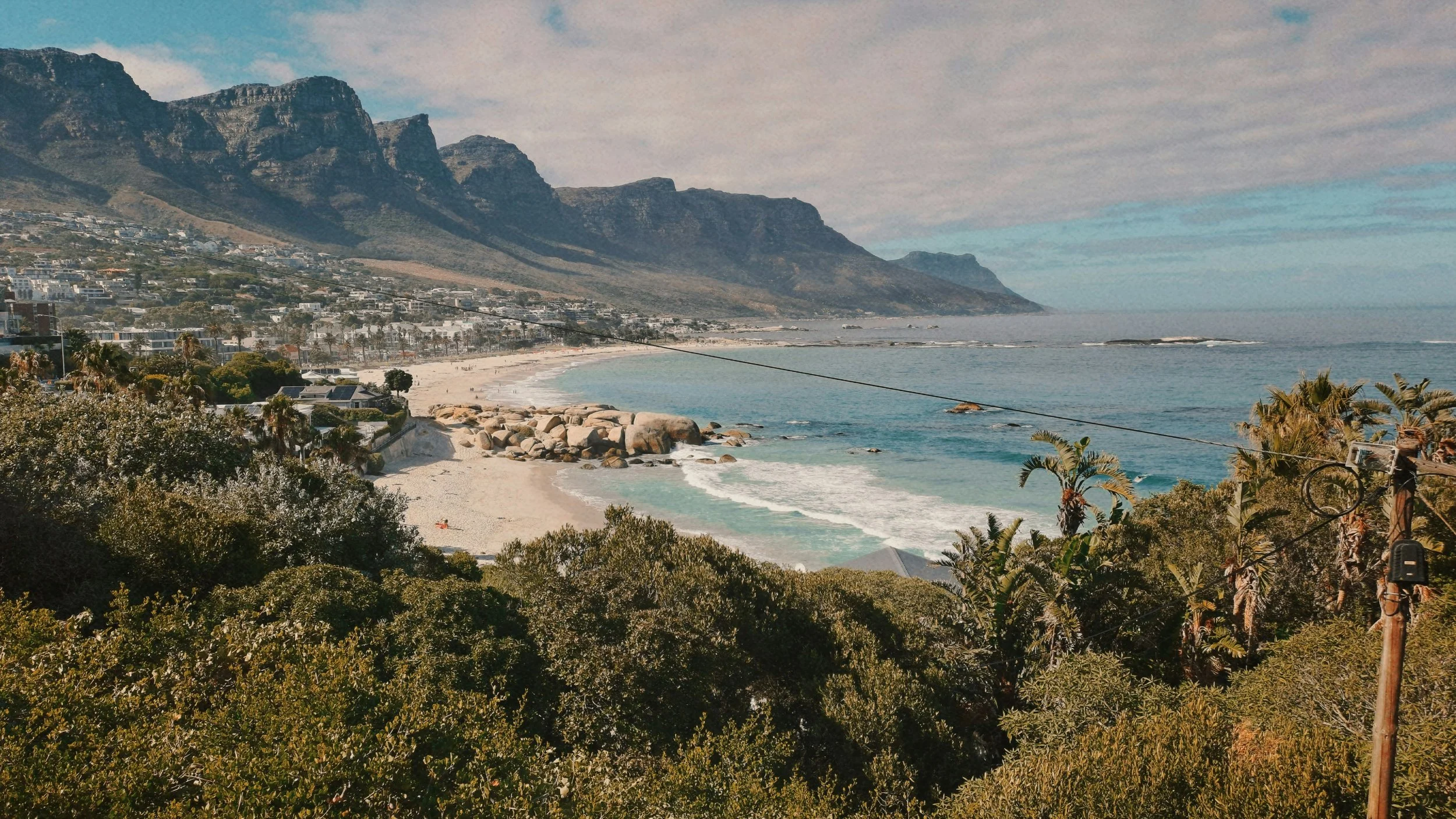 Coastal view of a beach with rocks, trees, and umbrellas, mountains in the background, and a partly cloudy sky.