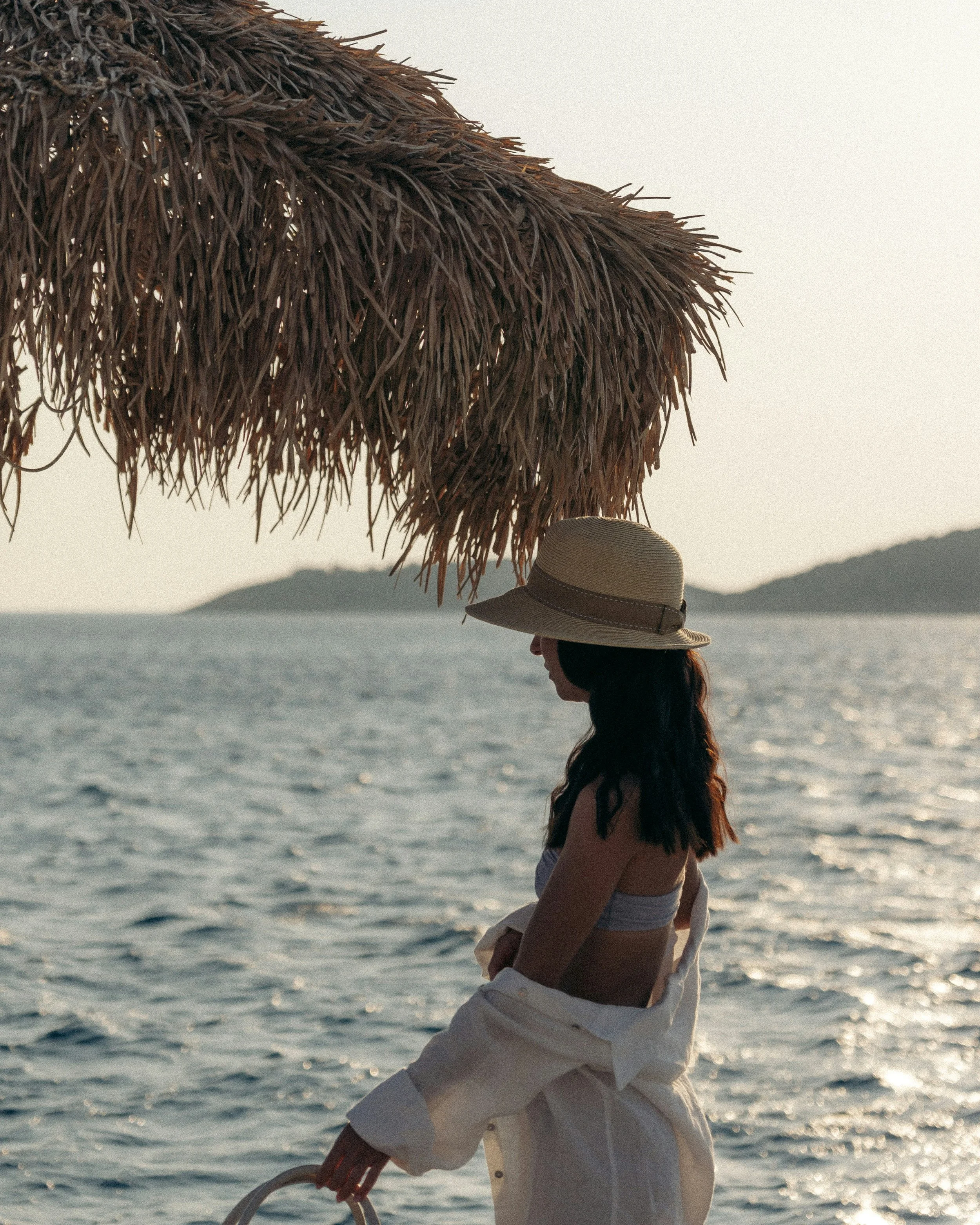 A woman in a straw hat and strapless dress stands on a beach near the water, holding a large thatched umbrella. The scene is during sunset, with calm waves and distant mountains in the background.