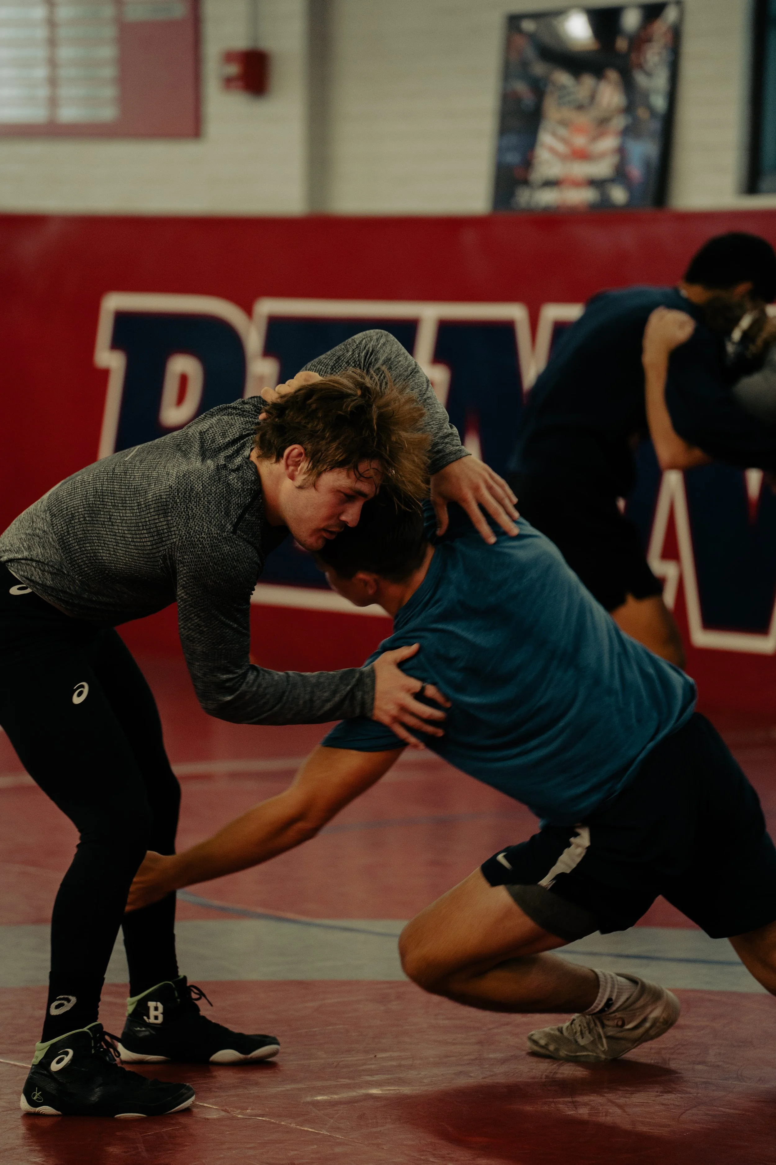 Two young men wrestling on a red wrestling mat, with one wearing a long-sleeved gray shirt and black sports shoes, and the other in a blue shirt and black shorts, in an indoor gym with a poster and a wall in the background.