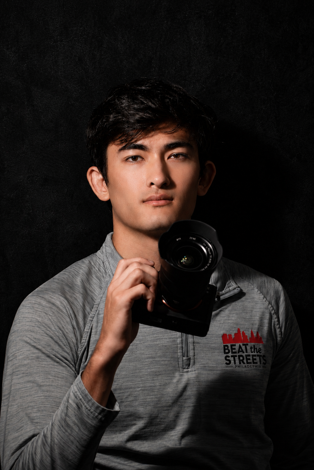 Young man in gray athletic top holding a camera against a black background.