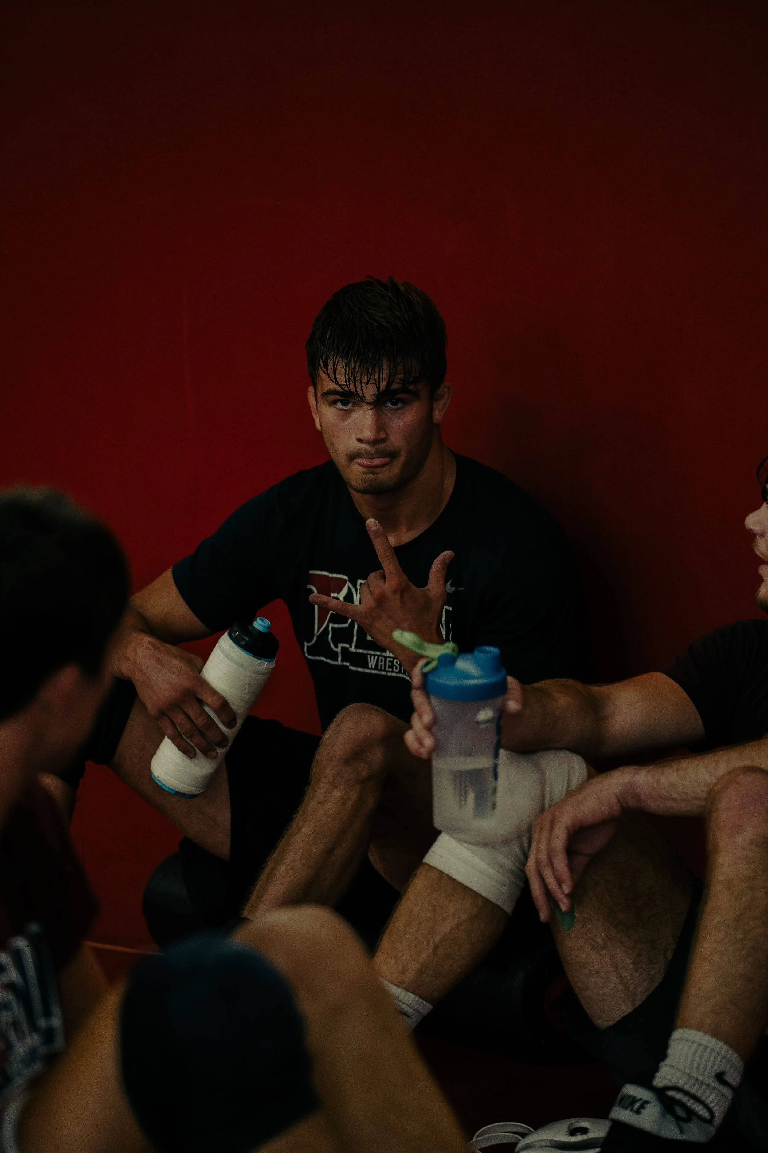 Group of wrestlers sitting together in a locker room, holding water bottles, with a red wall in the background.
