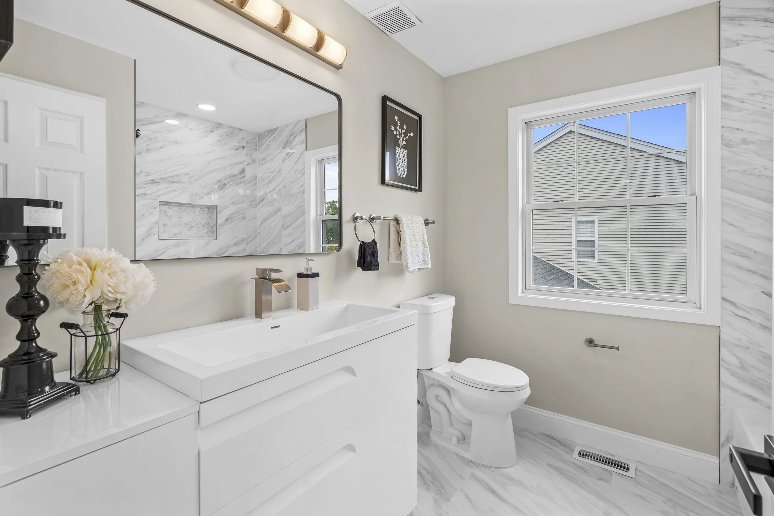 Bright modern bathroom with marble accents, white vanity with a sink, decorative items, towels, a large window, and a mirror.
