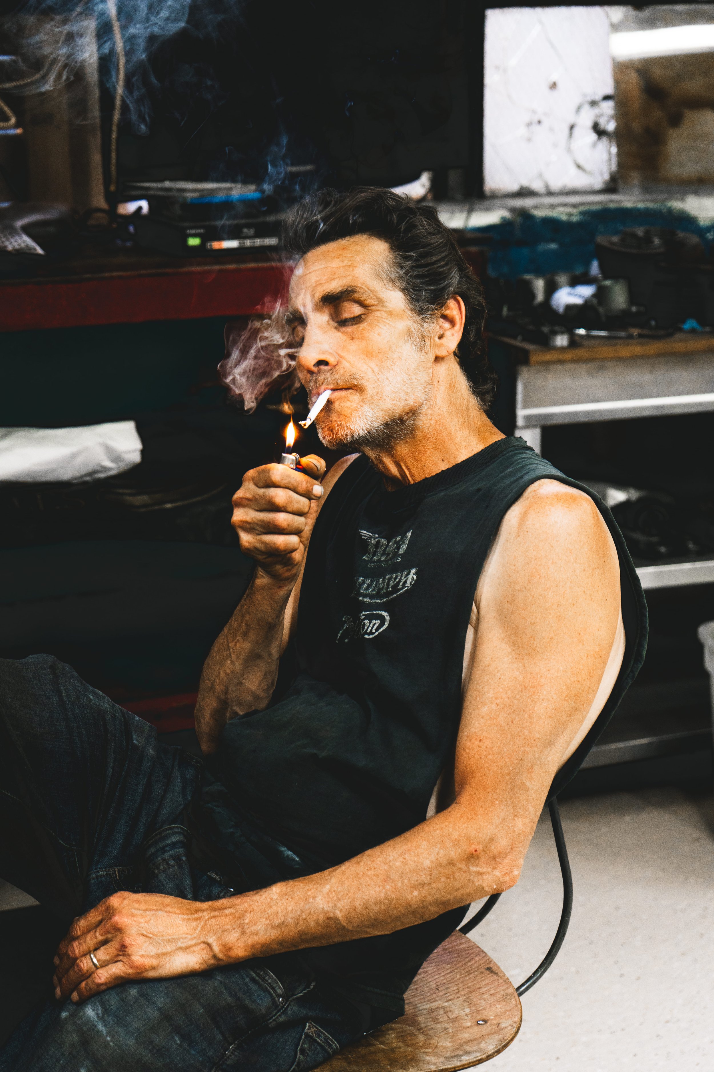 A man with gray hair and stubble, wearing a black sleeveless shirt and ripped jeans, sitting on a wooden chair, smoking a cigarette, with his eyes closed.