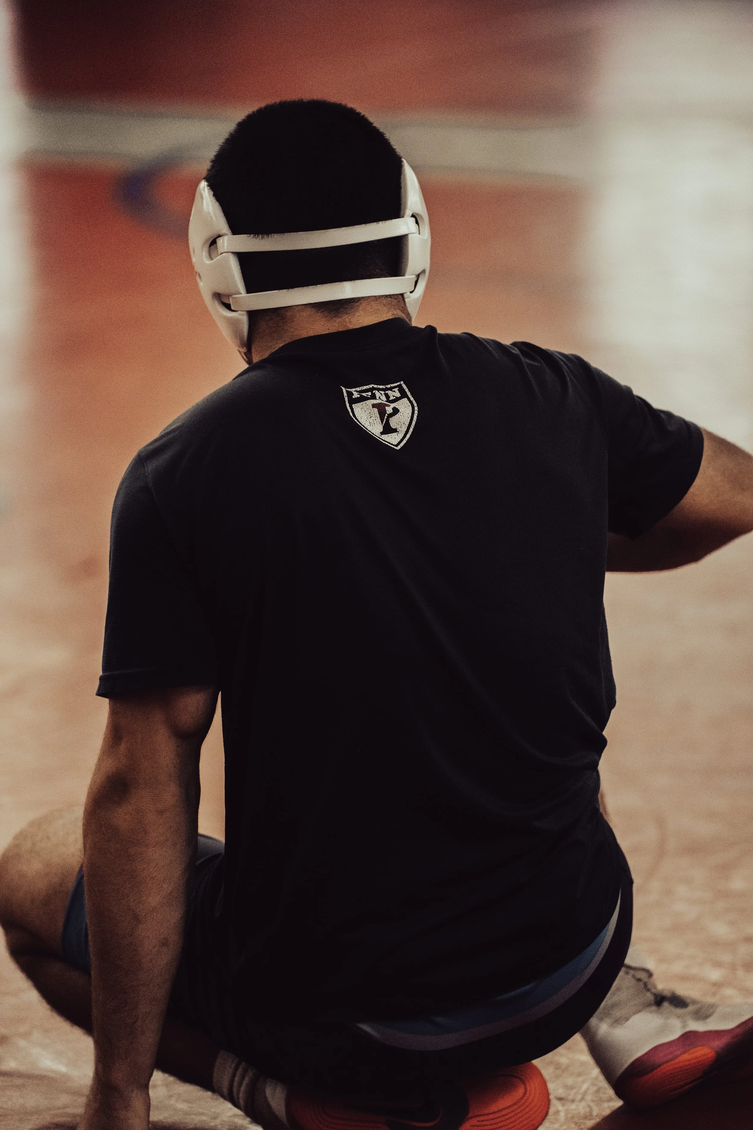 A person wearing a black shirt with a sports logo on the back, orange shoes, and a protective helmet, sitting on the ground.