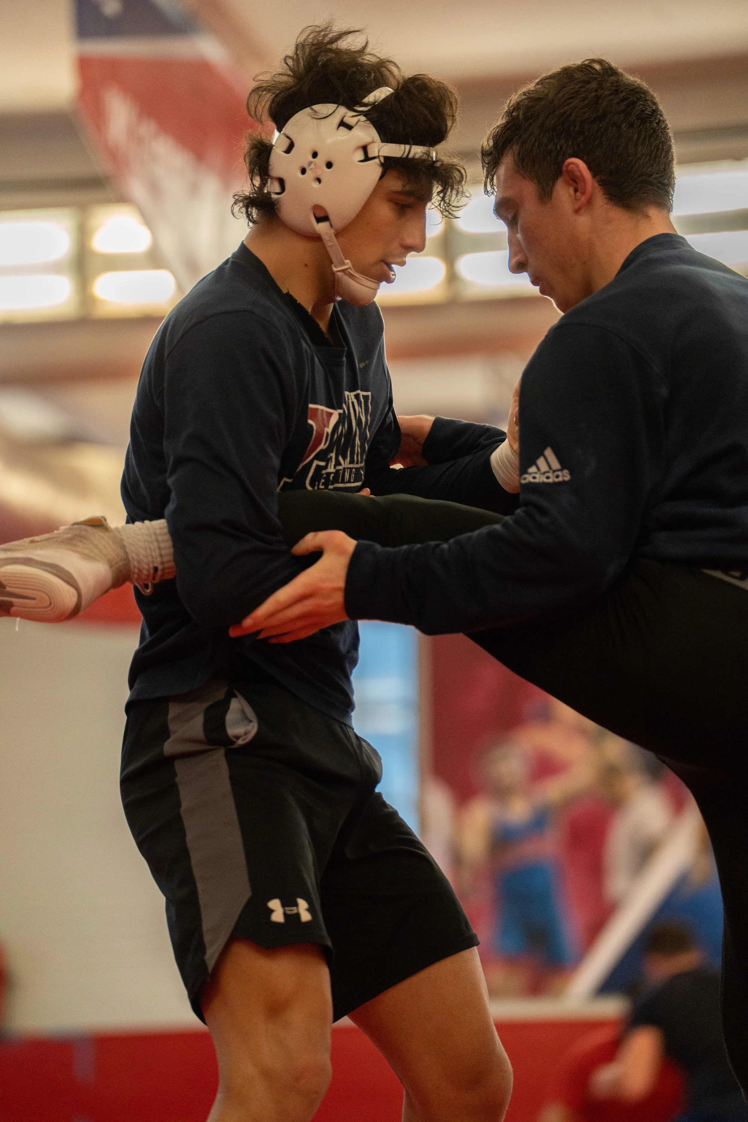 A male wrestler in black shorts and a navy shirt in a wrestling gym. He is being assisted by a coach or trainer holding him by the waist. The wrestler is wearing a protective headgear and has one leg raised. The background shows a gym with blurred pe