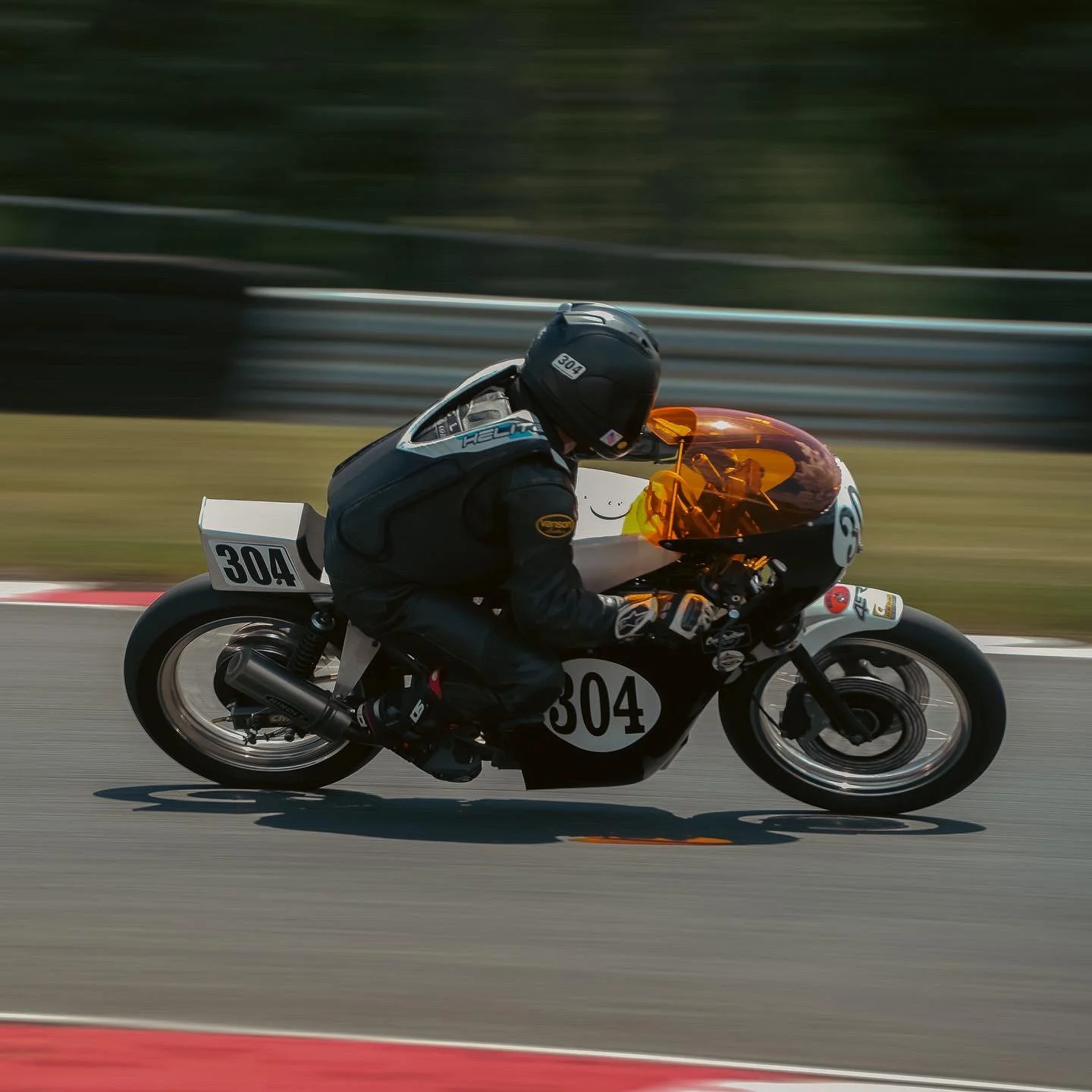 A motorcycle racer in black gear and helmet riding a black and white vintage racing motorcycle with number 304 on the side, on a race track with a blurred green background and guardrails.