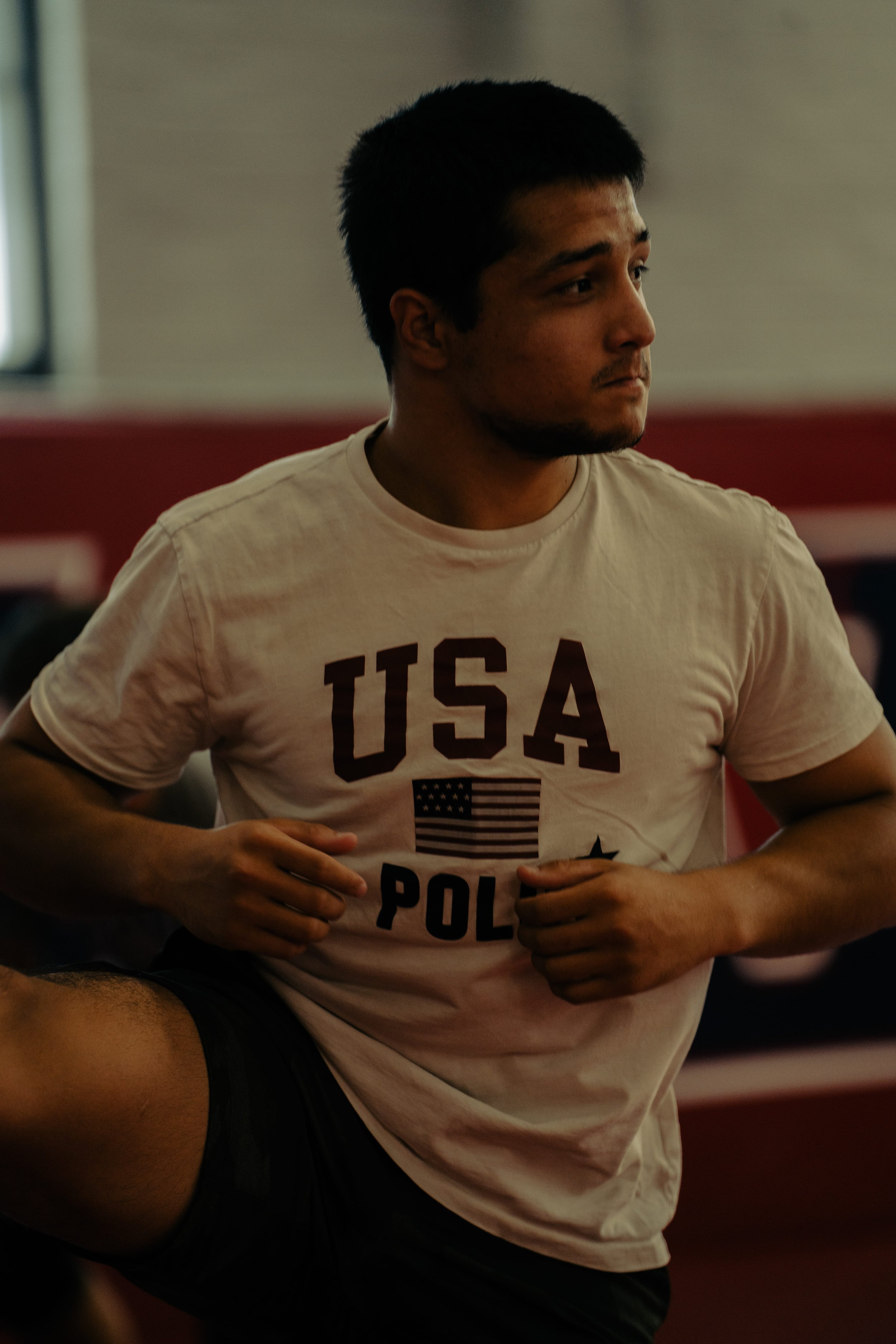 Young man with short dark hair and a beard, wearing a white T-shirt with 'USA POL' and American flag, in a gym or training environment.