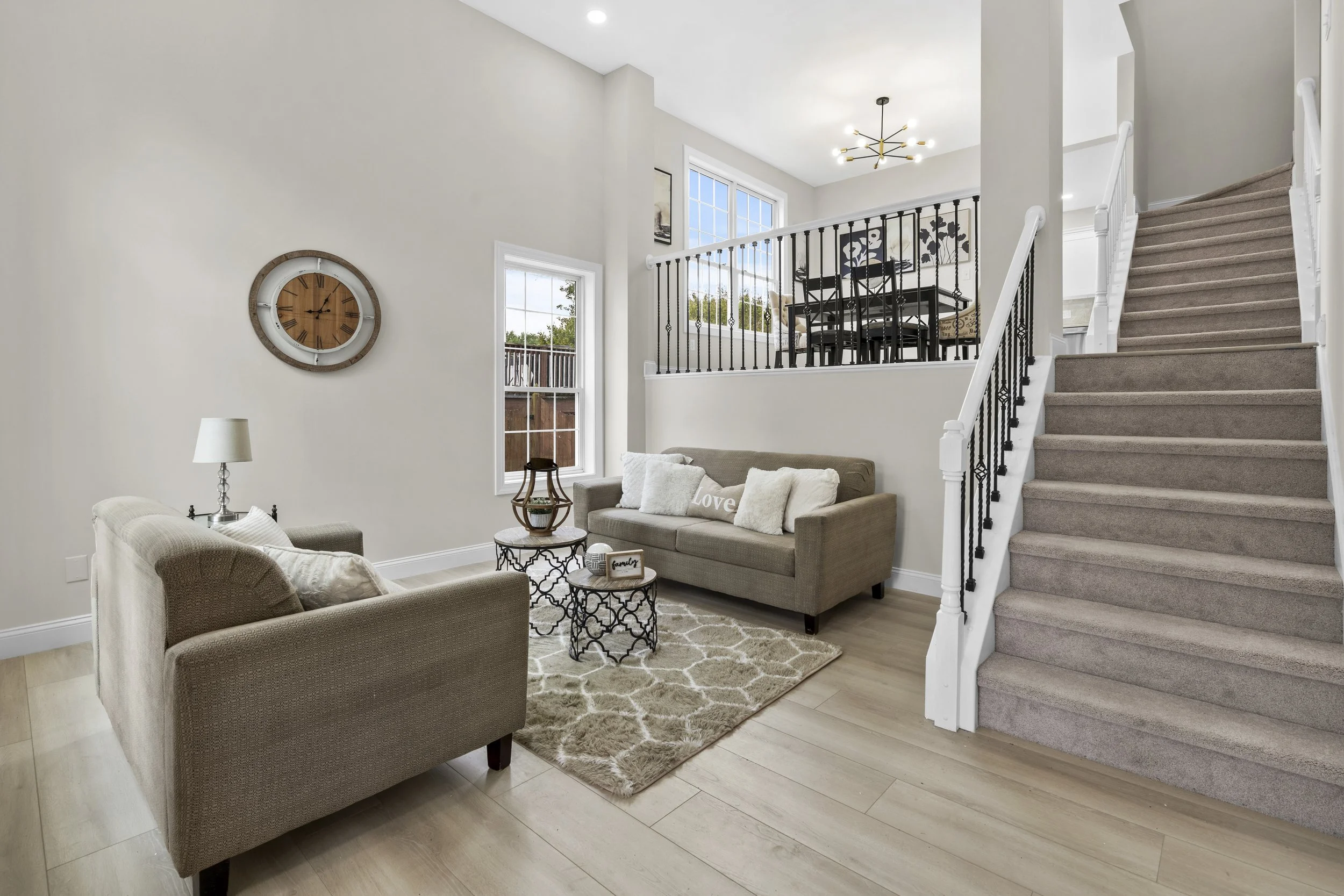 Living room with beige sofas, a coffee table, a rug, wall clock, lamps, and a staircase with carpeted steps and a black railing, illuminated by natural light from multiple large windows.