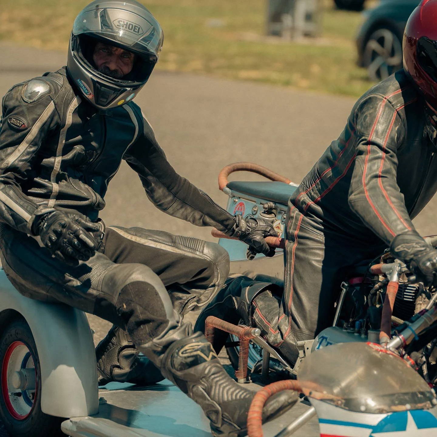 Two men dressed in motorcycle gear, one wearing a black helmet and the other wearing a red helmet, sitting on a motorcycle with a sidecar during daytime.