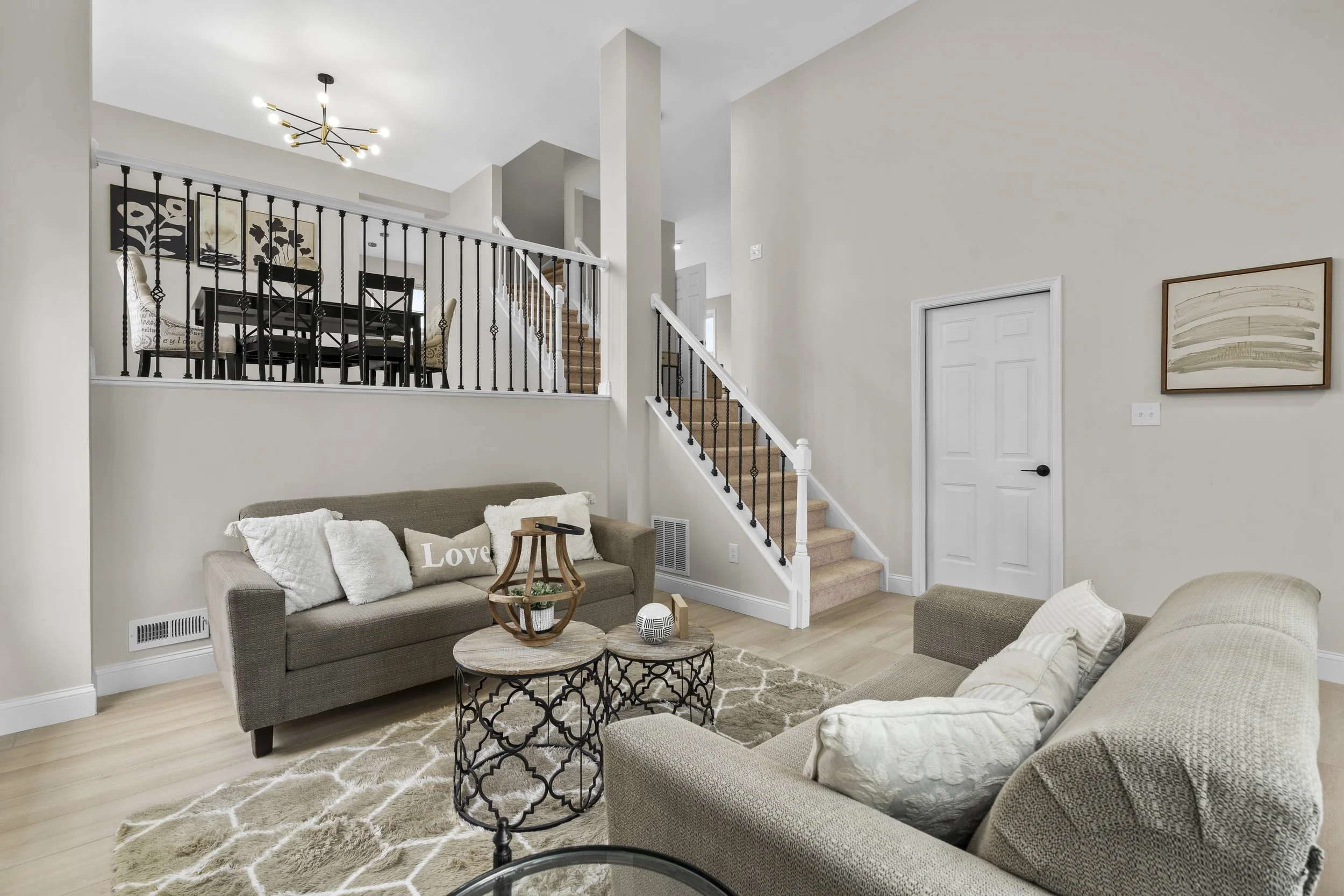 Living room with beige sofas, a patterned rug, and a marble-top coffee table. Walls have artwork, stairs with black iron railing lead to an upper level, and a dining area with a black table and chairs is visible in the background.
